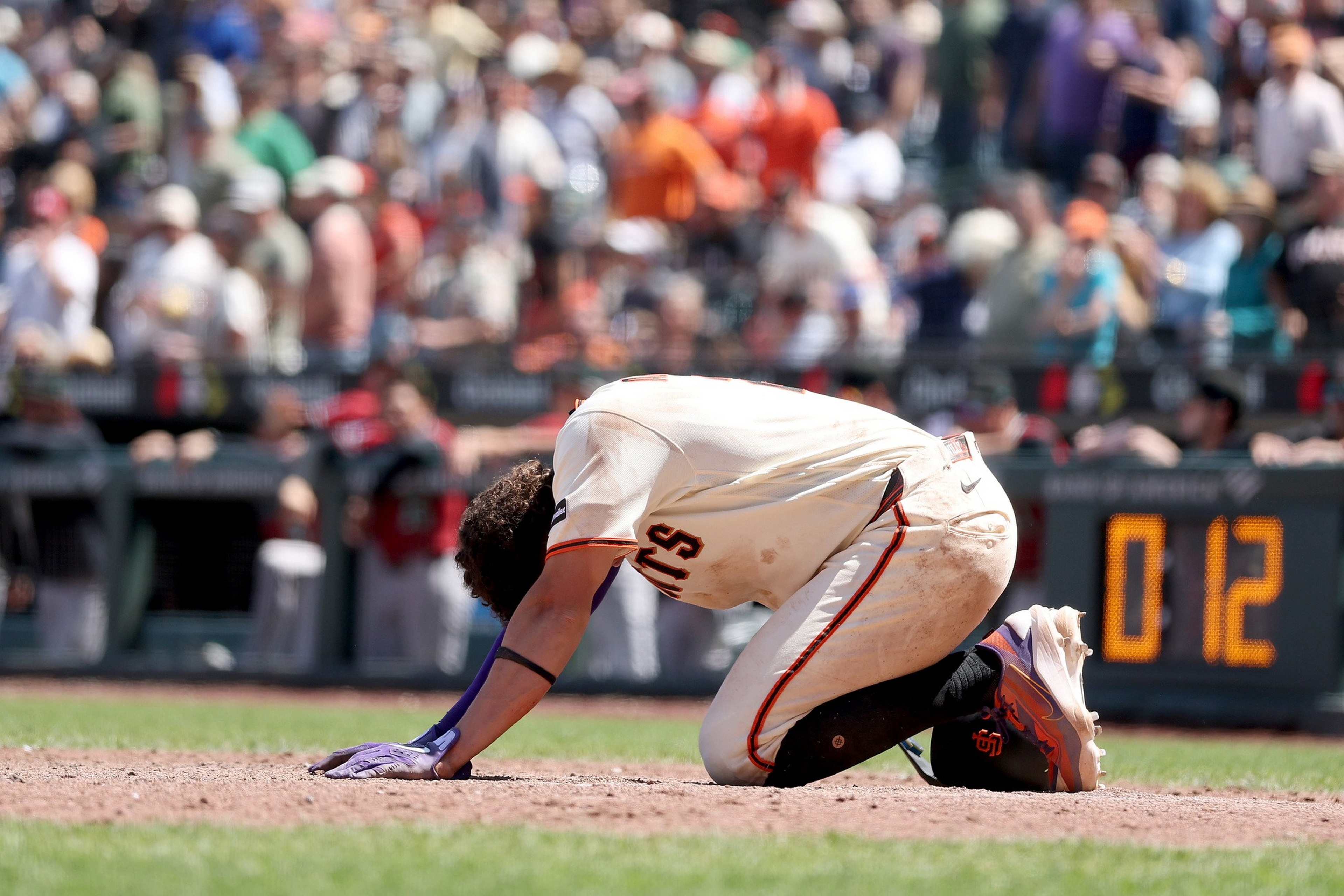 A baseball player kneels on the ground with head bowed and one hand touching the dirt, while a crowd watches in the background.