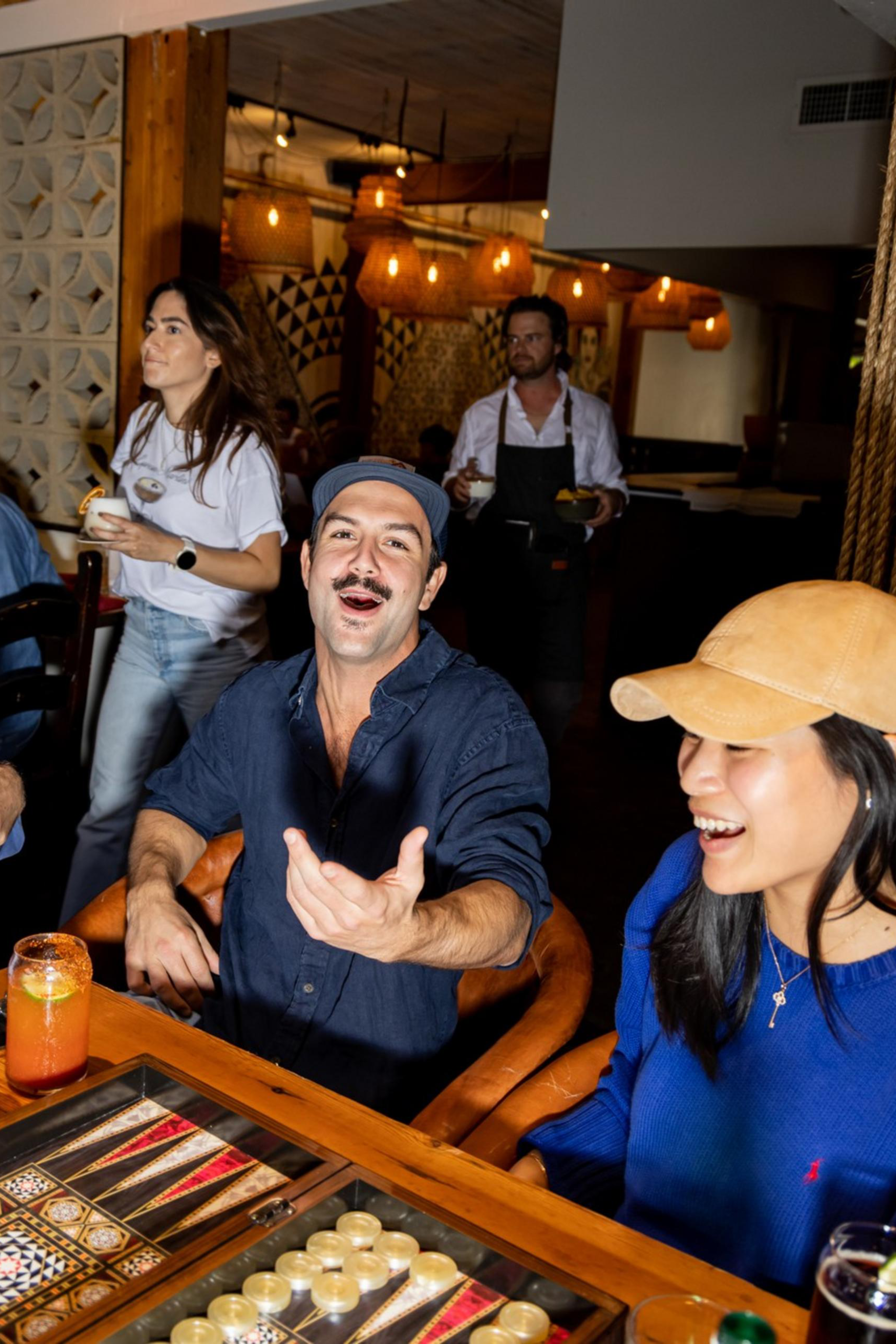 Three people are playing backgammon at a table with drinks, laughing and gesturing, while others stand or walk in the warmly lit room behind them.