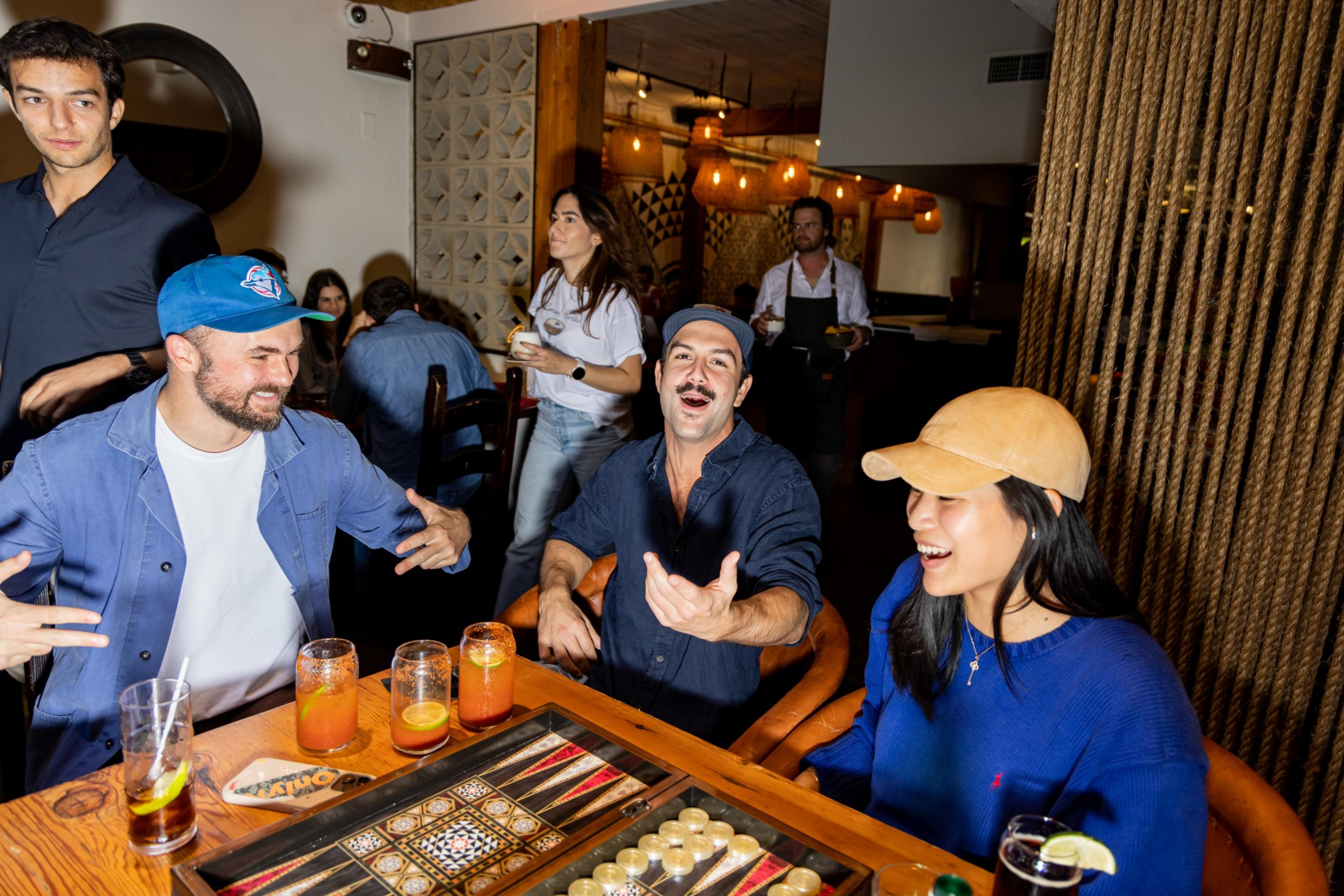 Three people are playing backgammon at a table with drinks, laughing and gesturing, while others stand or walk in the warmly lit room behind them.