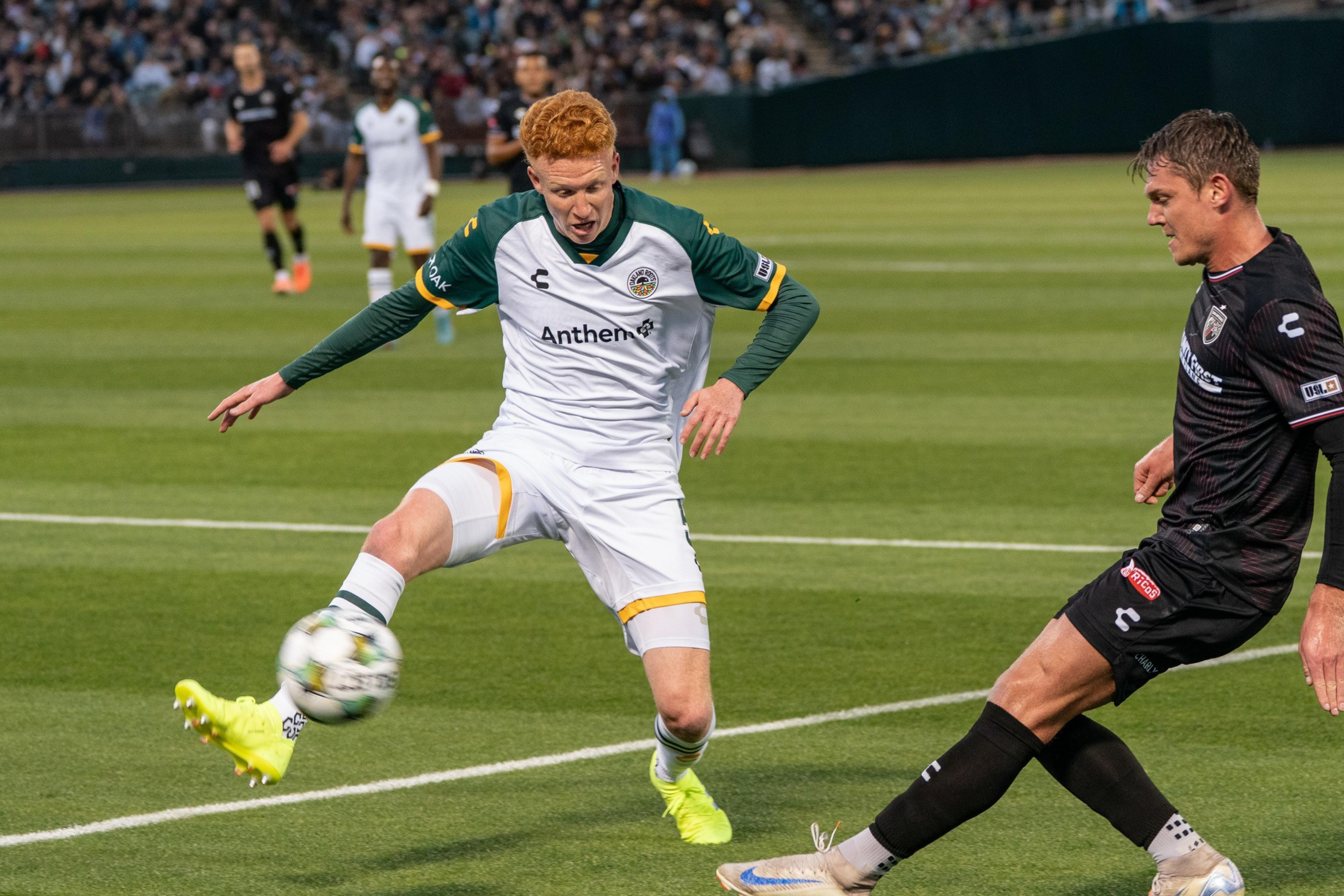 A soccer player in a white and green jersey skillfully kicks a ball mid-air, while another player in black defends. The crowd watches from the stands.