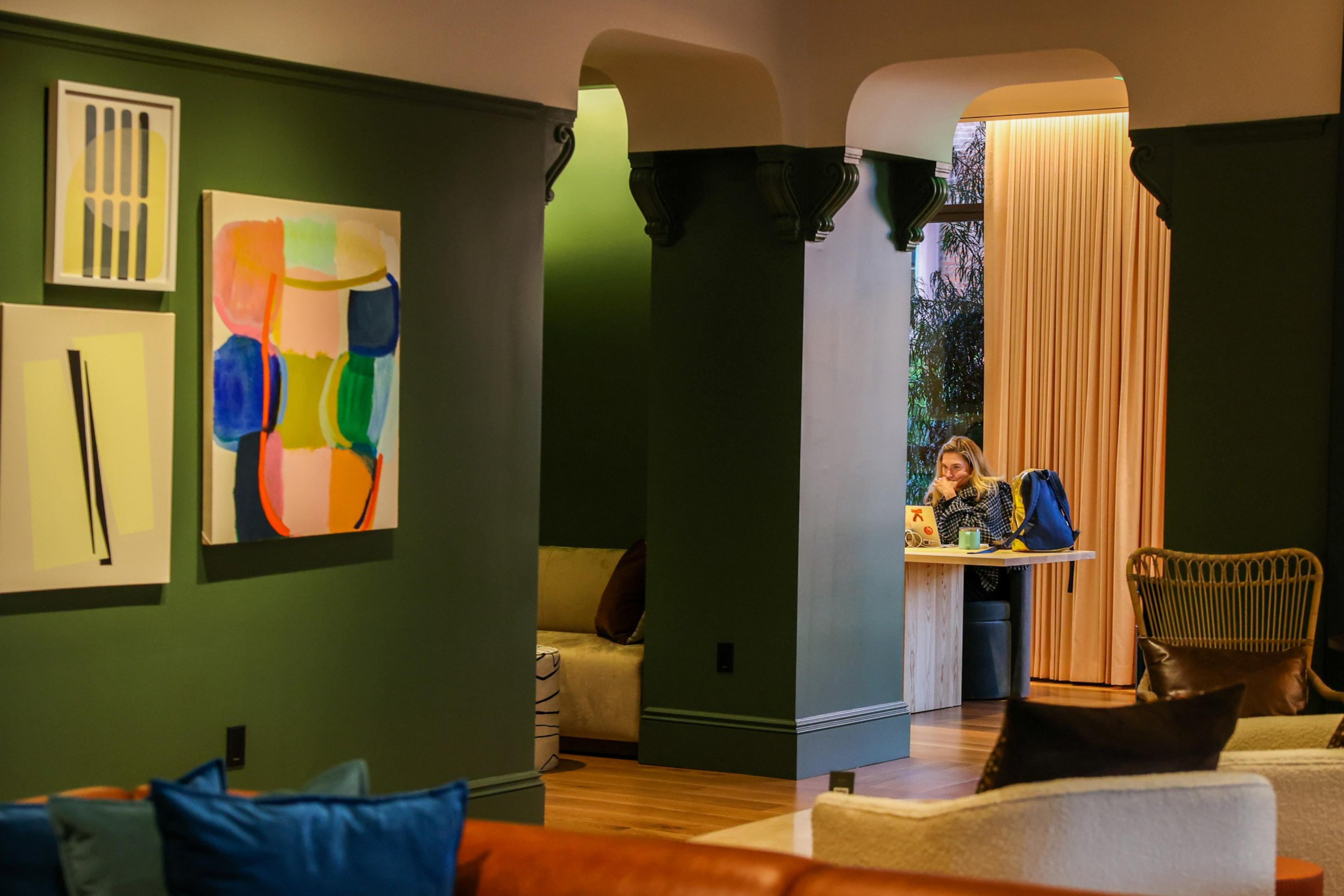 A person sits at a small table with a laptop and a coffee cup in a cozy room with green walls, colorful abstract art, and cushioned seating.