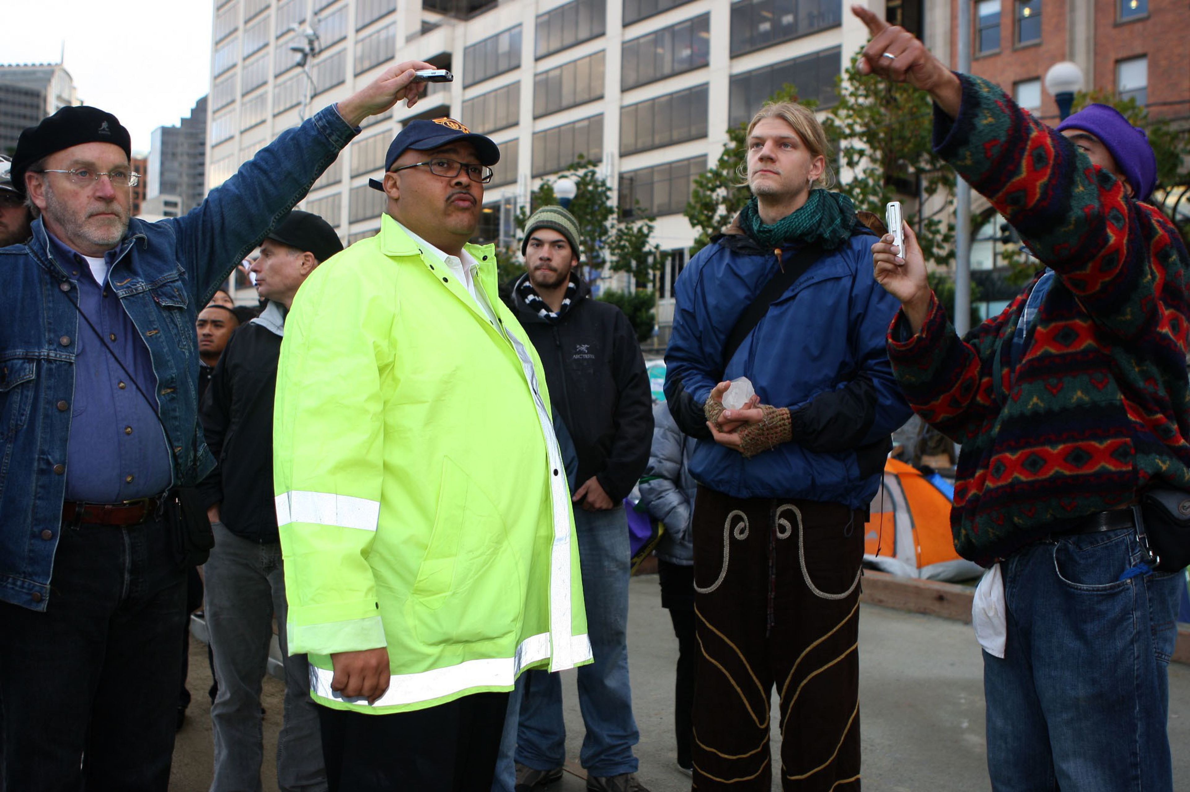Several people stand outdoors in an urban area; one in a bright yellow jacket, others in layered jackets, two holding small devices to record or measure.