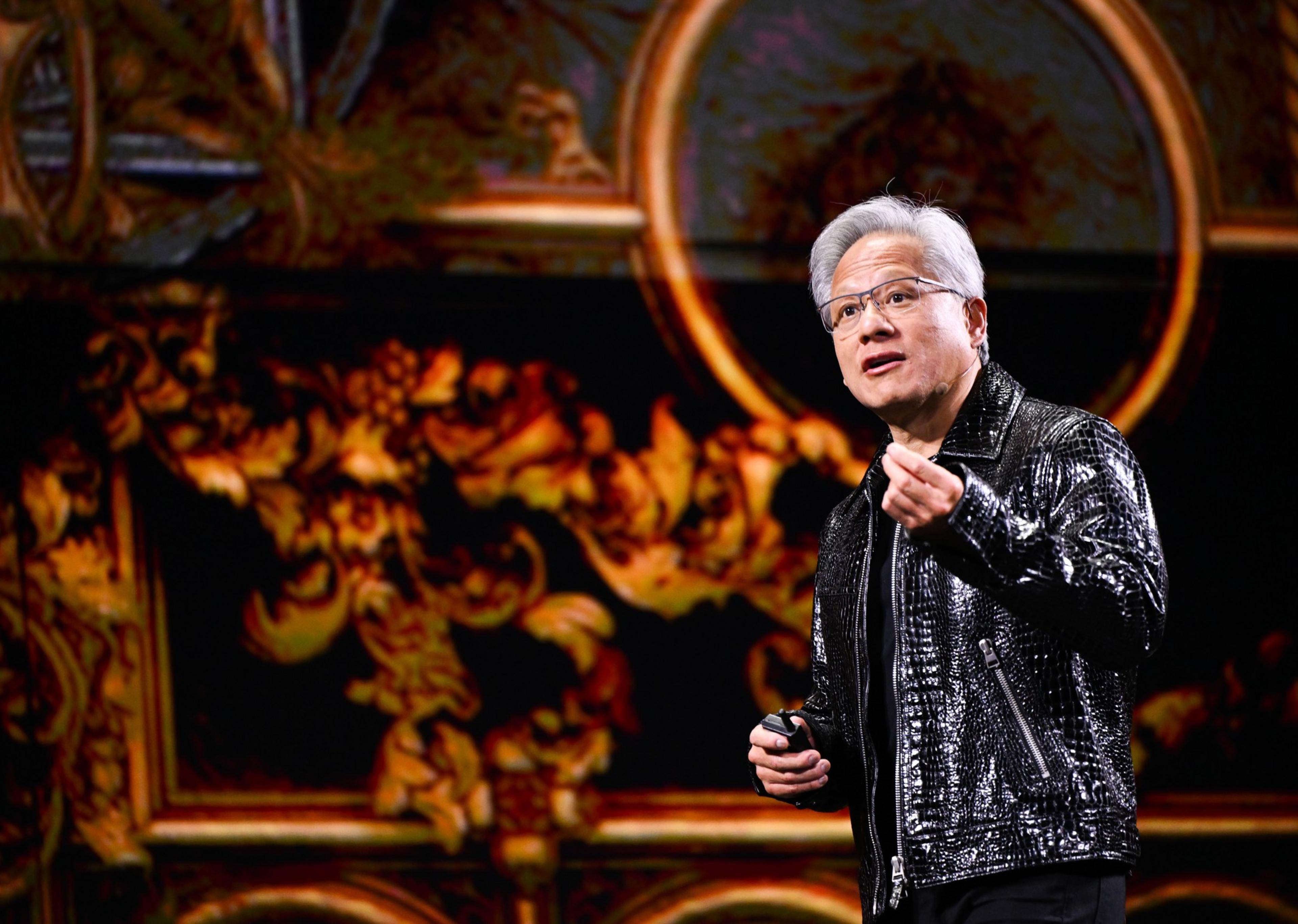 A man with white hair and glasses wearing a shiny black jacket gestures while speaking in front of a detailed, ornate backdrop with dark and gold tones.