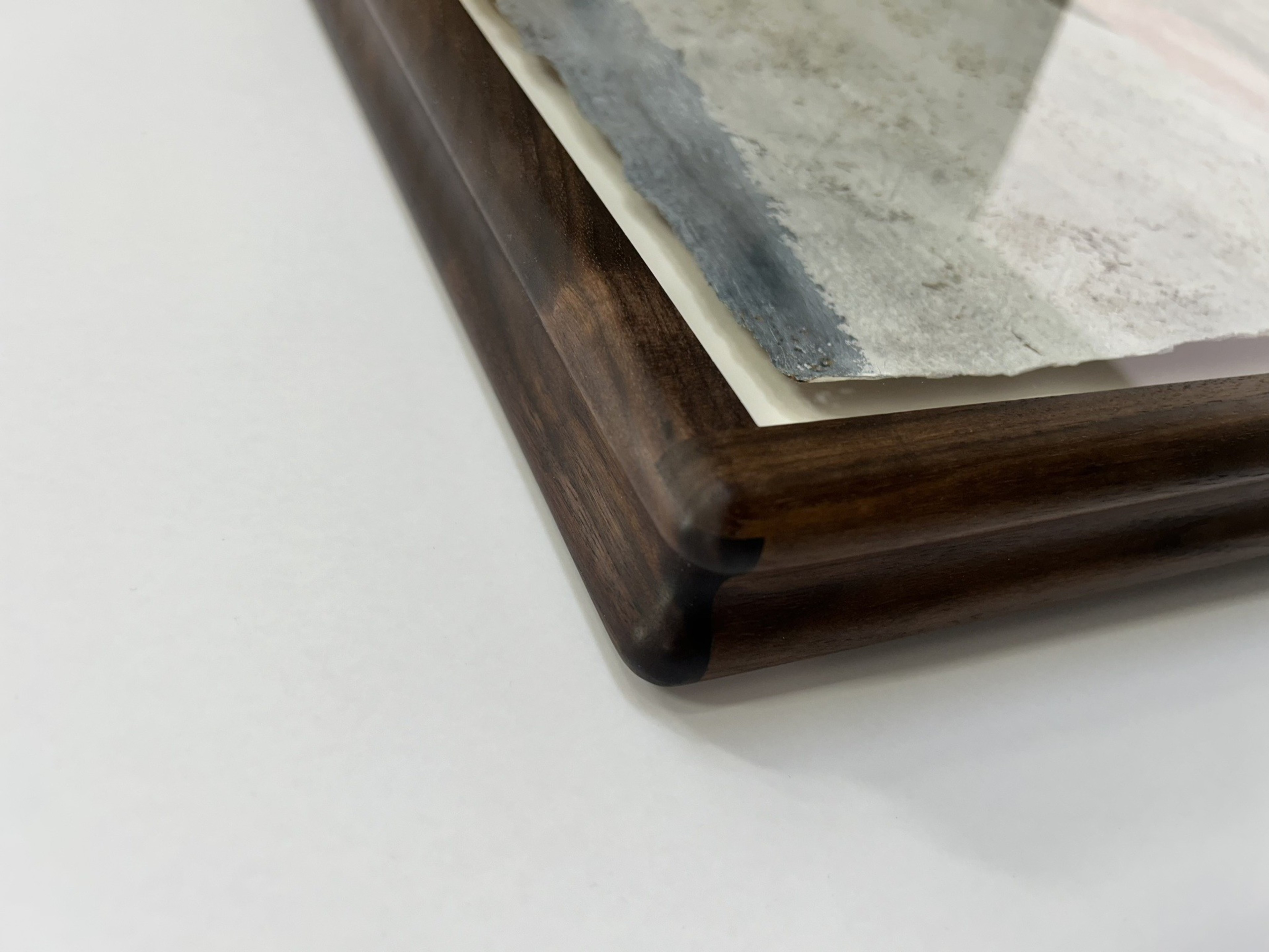 A close-up corner of a wooden picture frame with dark finish, holding a textured, weathered paper artwork.
