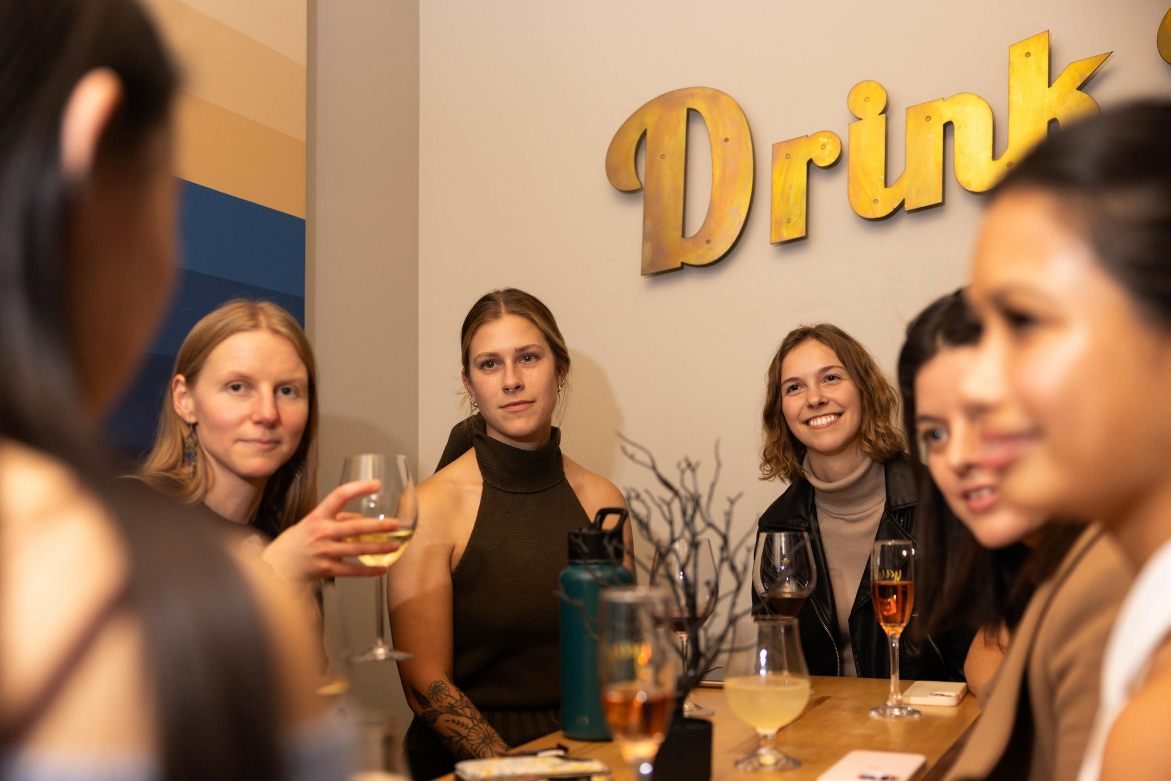 A group of women holding glasses of wine sit around a table in a bar listening attentively to a person off camera.