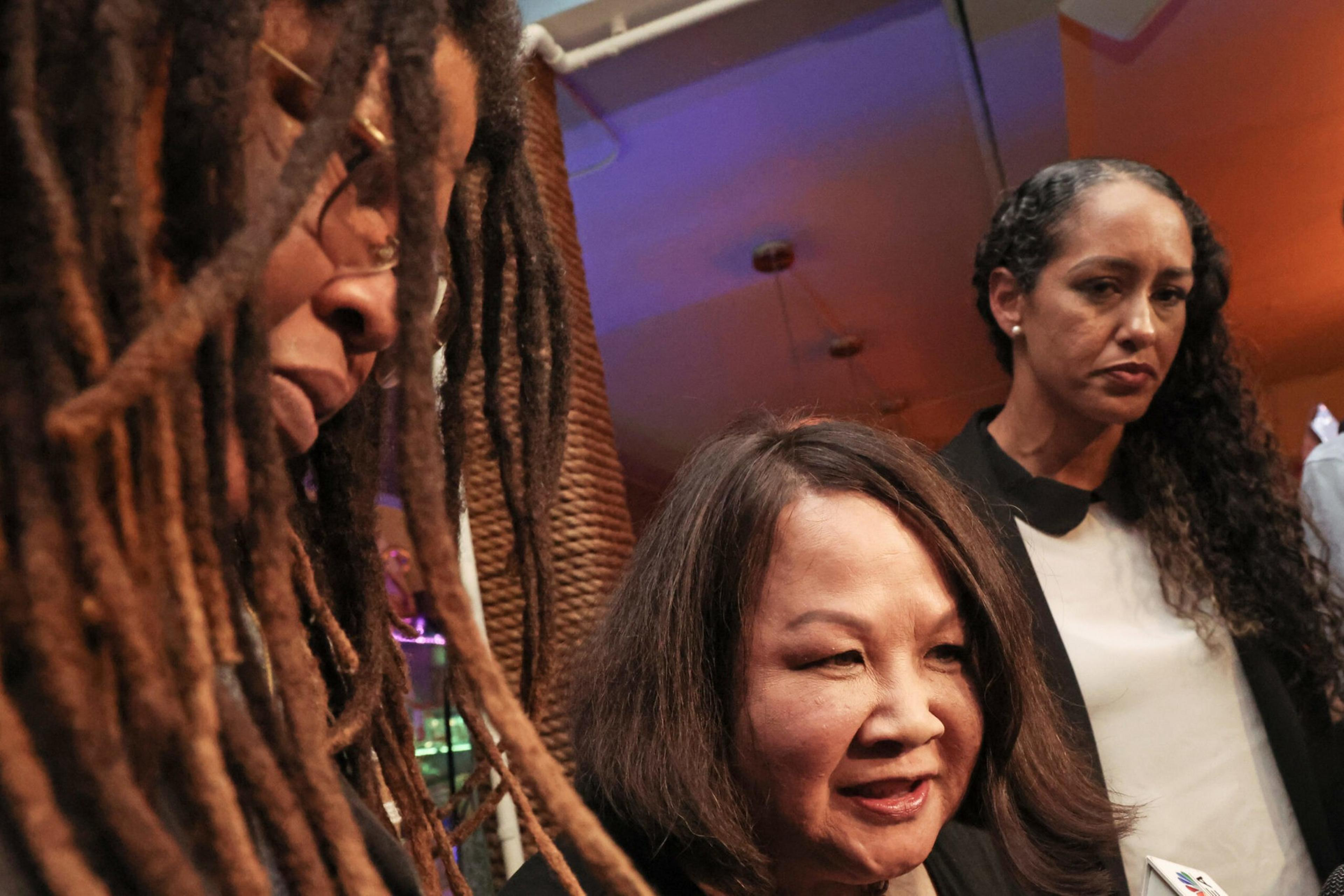 Three women are closely gathered indoors; one with long dreadlocks, one smiling with straight hair, and one serious-looking with curly hair.