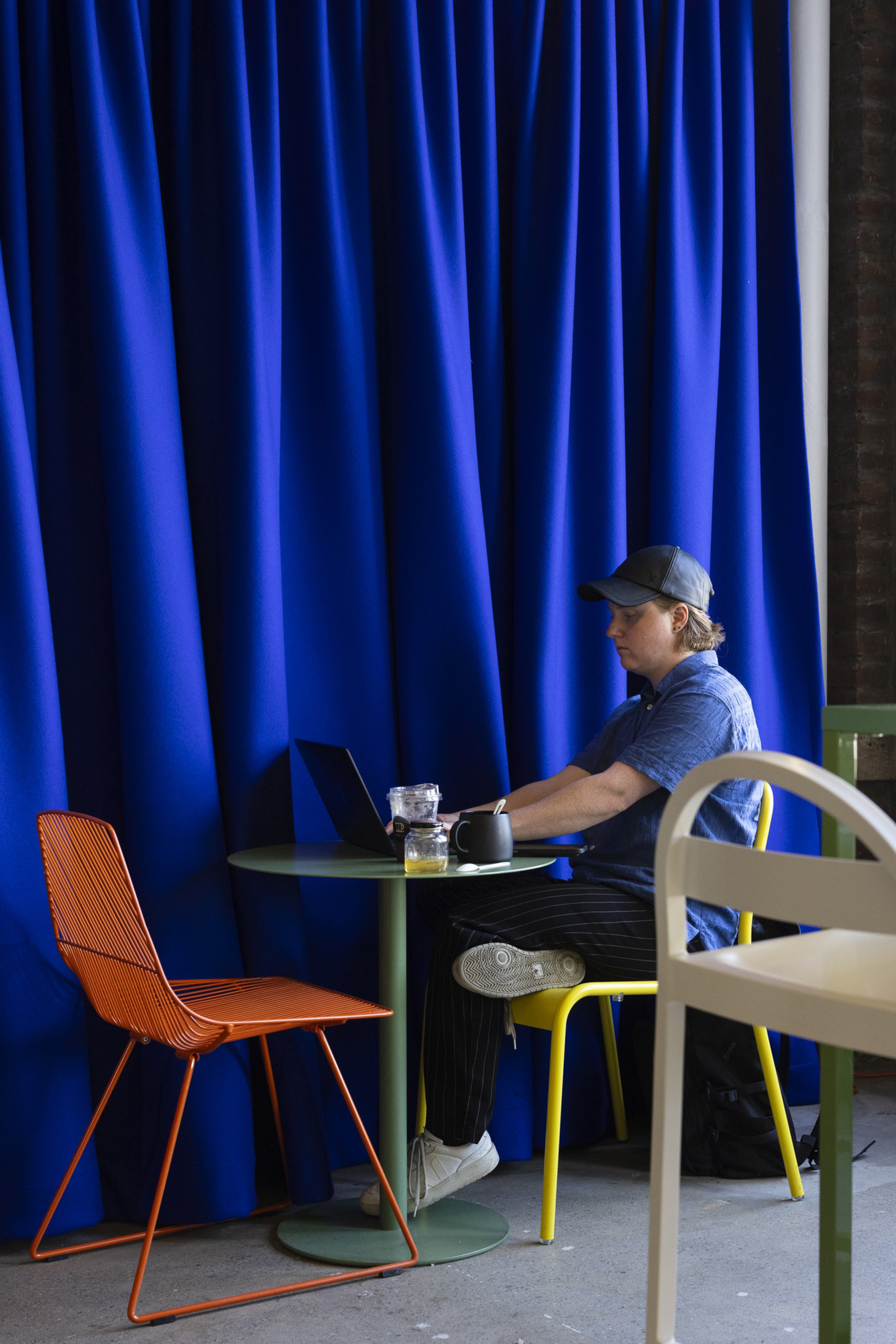 A person wearing a blue shirt and cap sits on a yellow chair at a small round green table, working on a laptop with a drink nearby and an empty orange chair opposite.