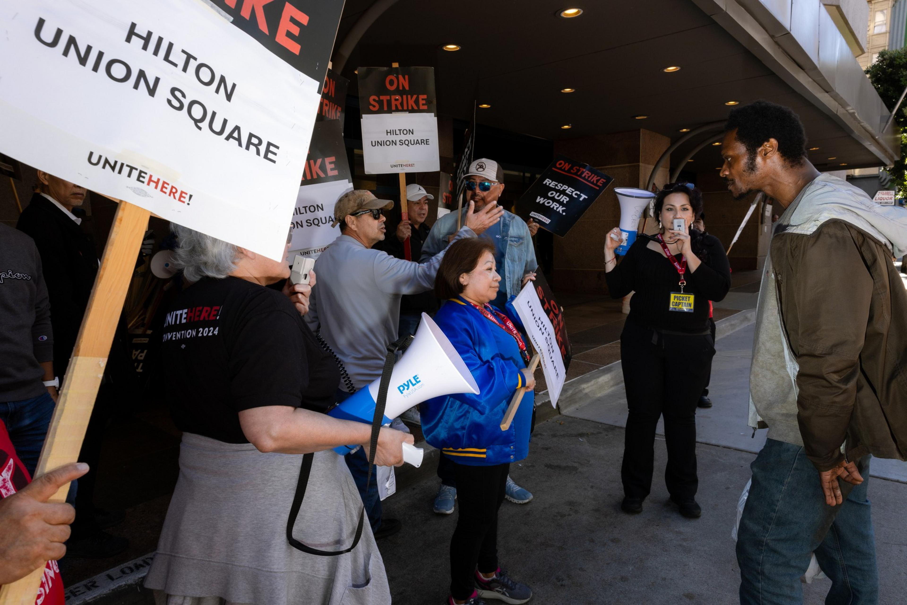 A group of people, holding signs reading &quot;ON STRIKE&quot; and &quot;Hilton Union Square,&quot; gathered for a protest. Some have megaphones, engaging with a passerby.