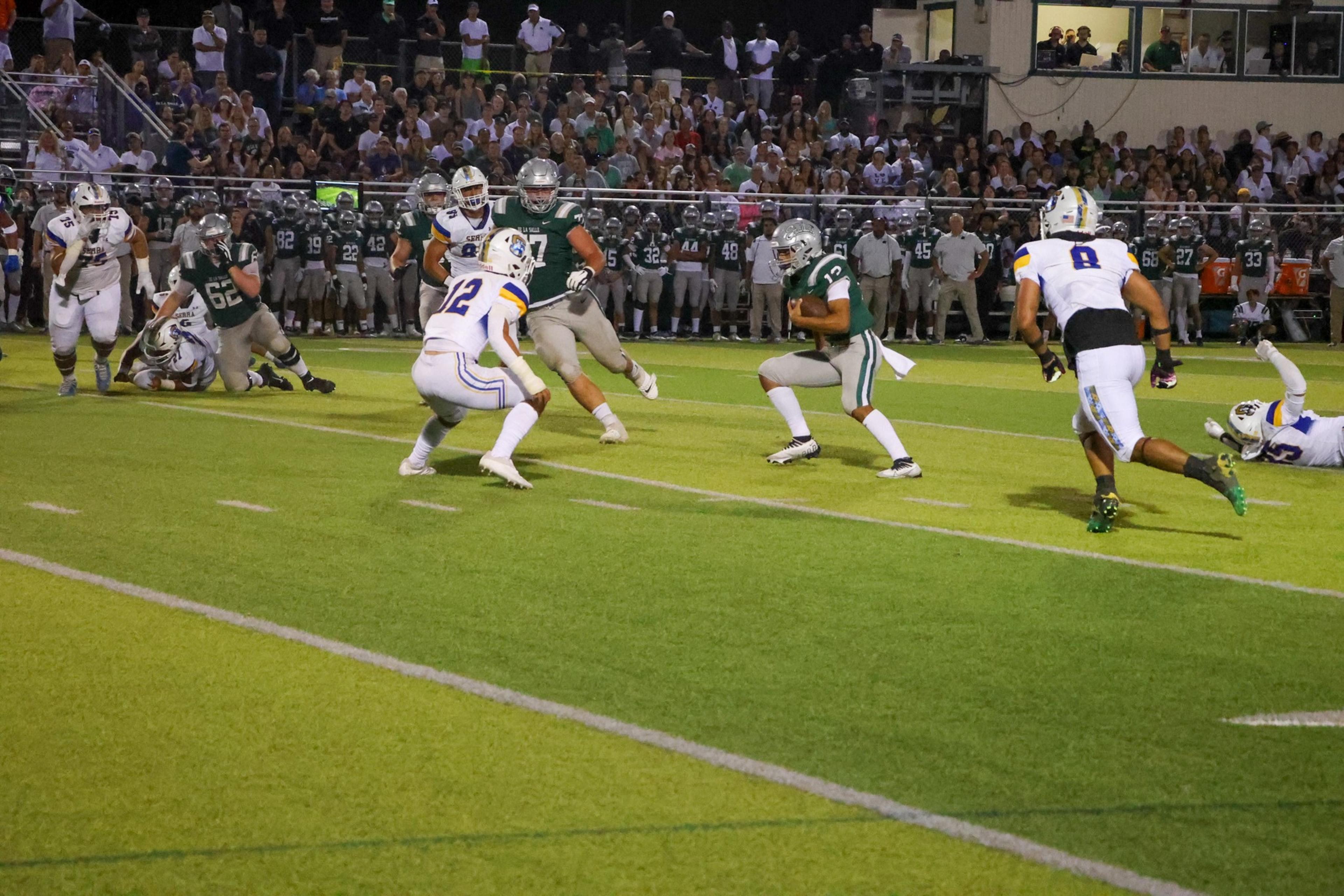 A football player in green runs with the ball as opponents in white approach, while teammates and spectators watch from the sidelines.
