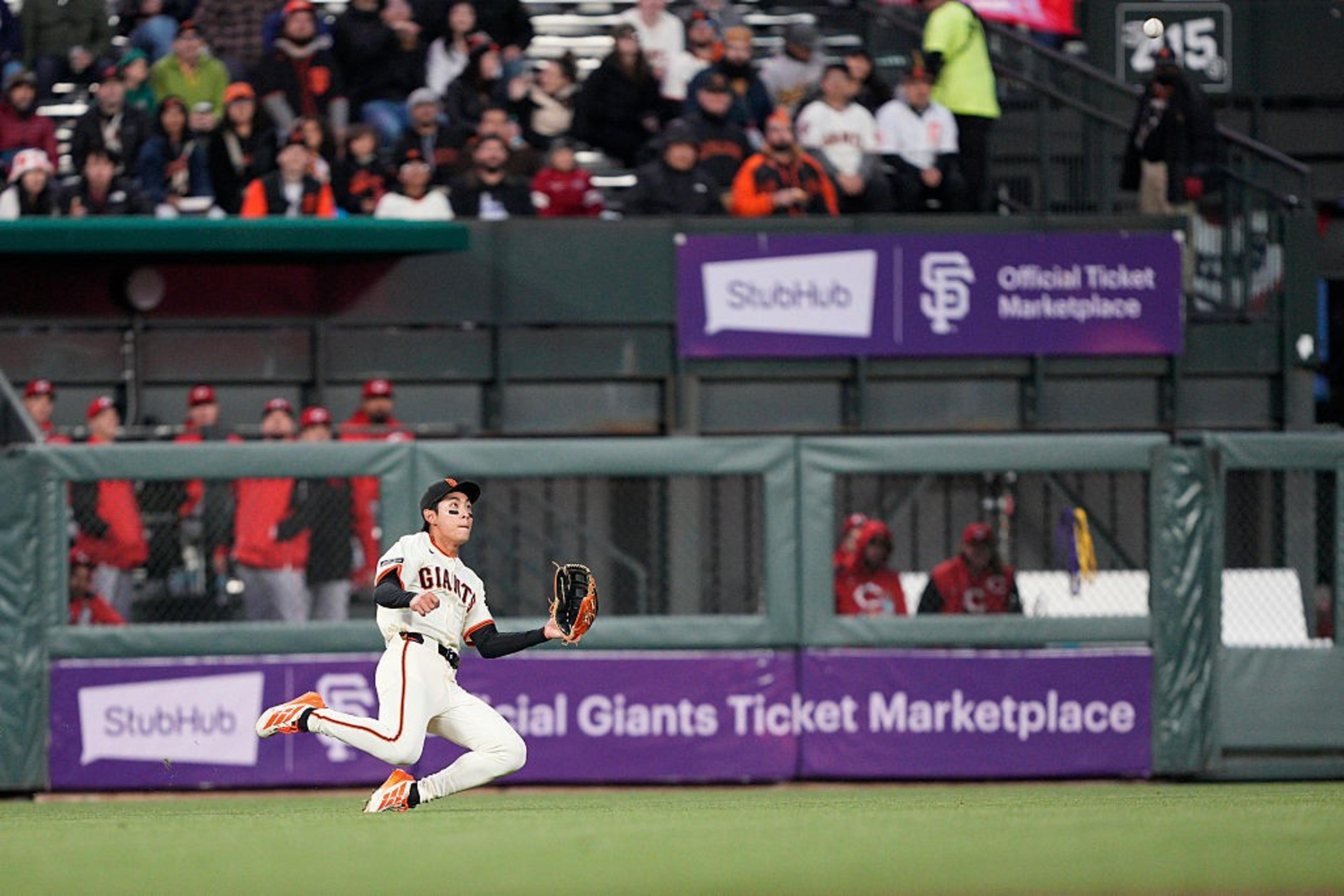 A baseball player in a Giants uniform is mid-action with a glove extended to catch a ball on a field. Spectators and team members are visible in the background.