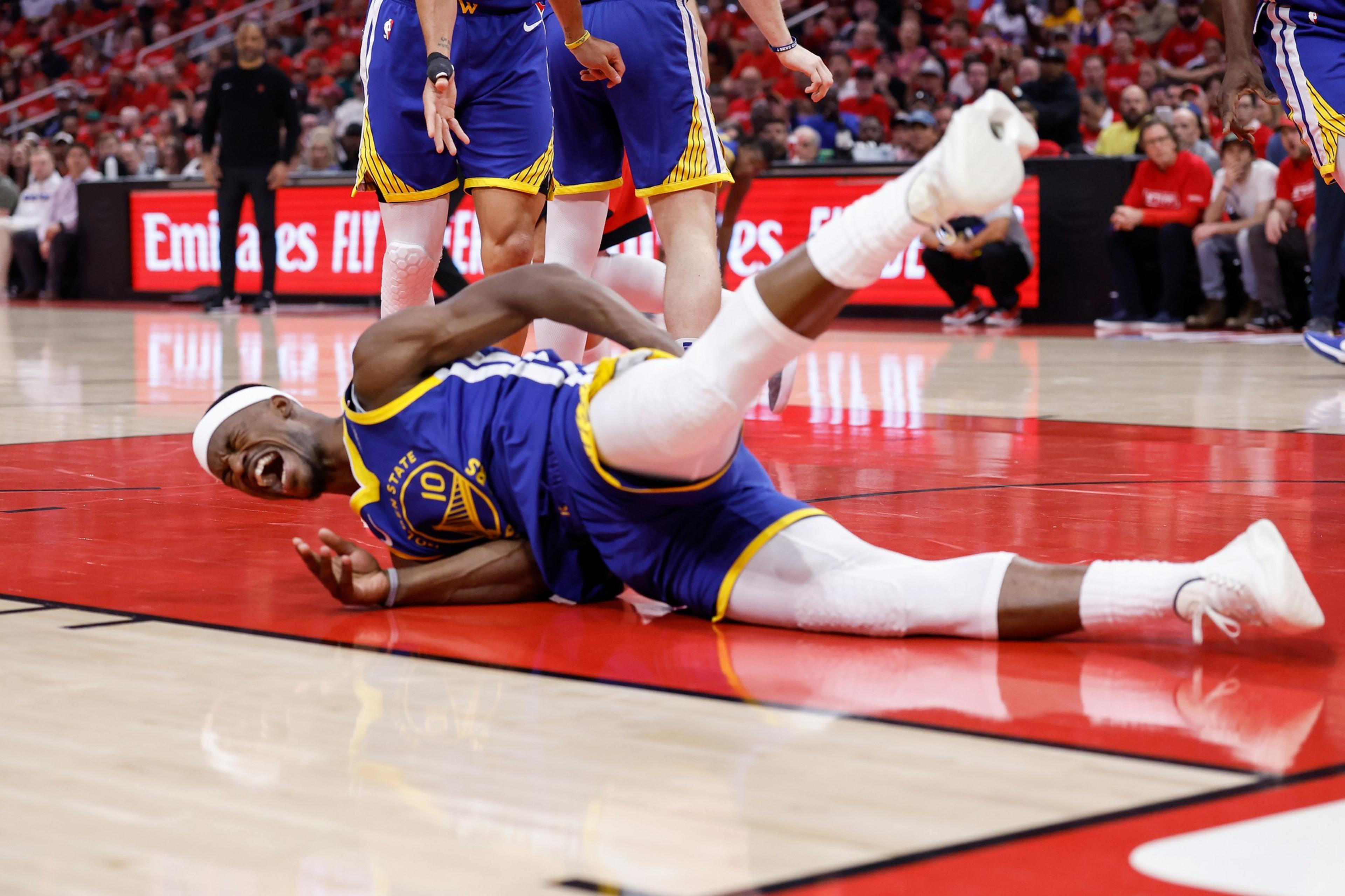 A basketball player in a blue and yellow uniform lies on the court in pain, holding his side. Teammates stand nearby, and spectators watch in a crowded arena.