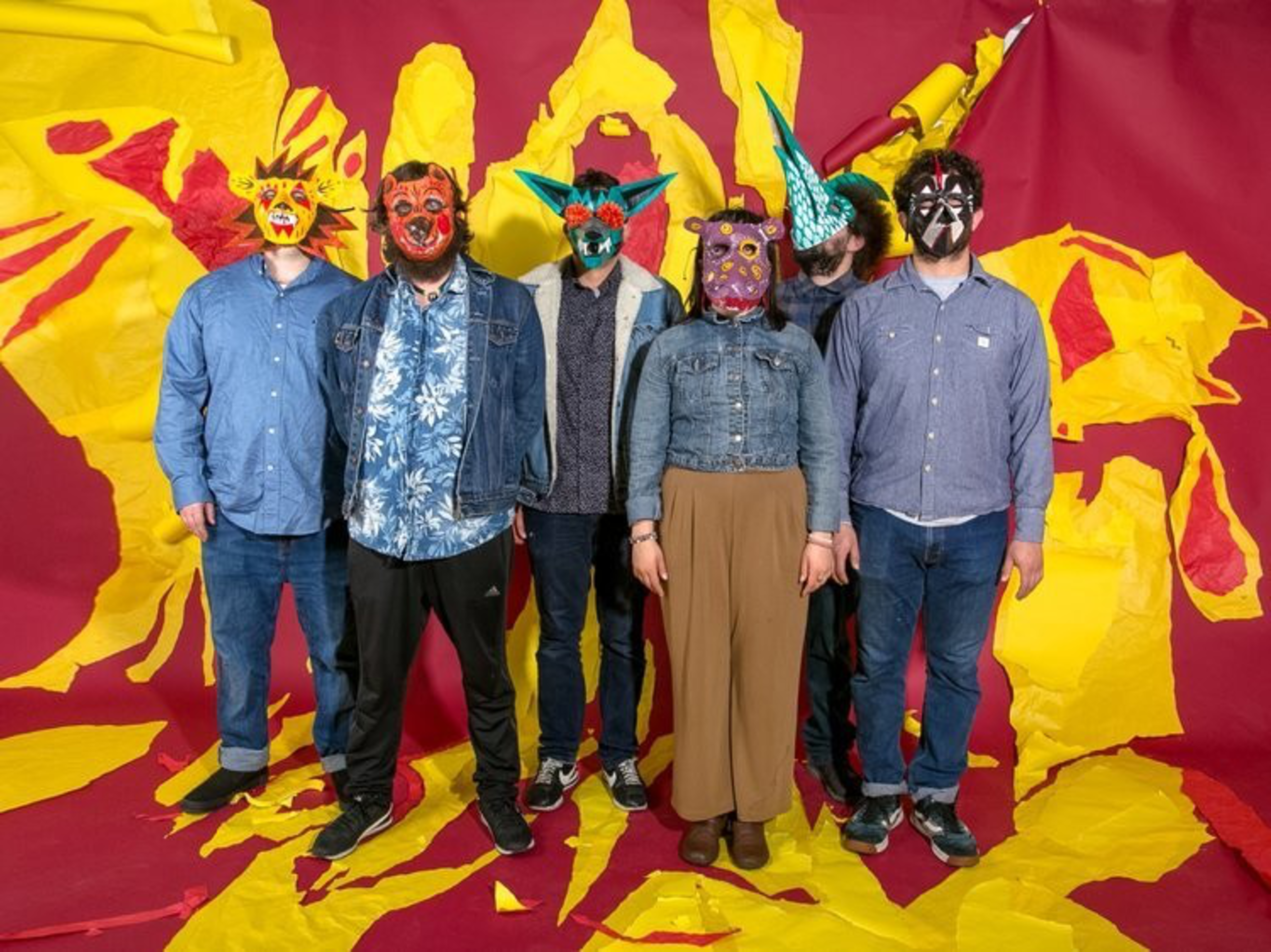 Six people stand against a red and yellow torn paper backdrop, each wearing colorful, animalistic masks and casual clothing.