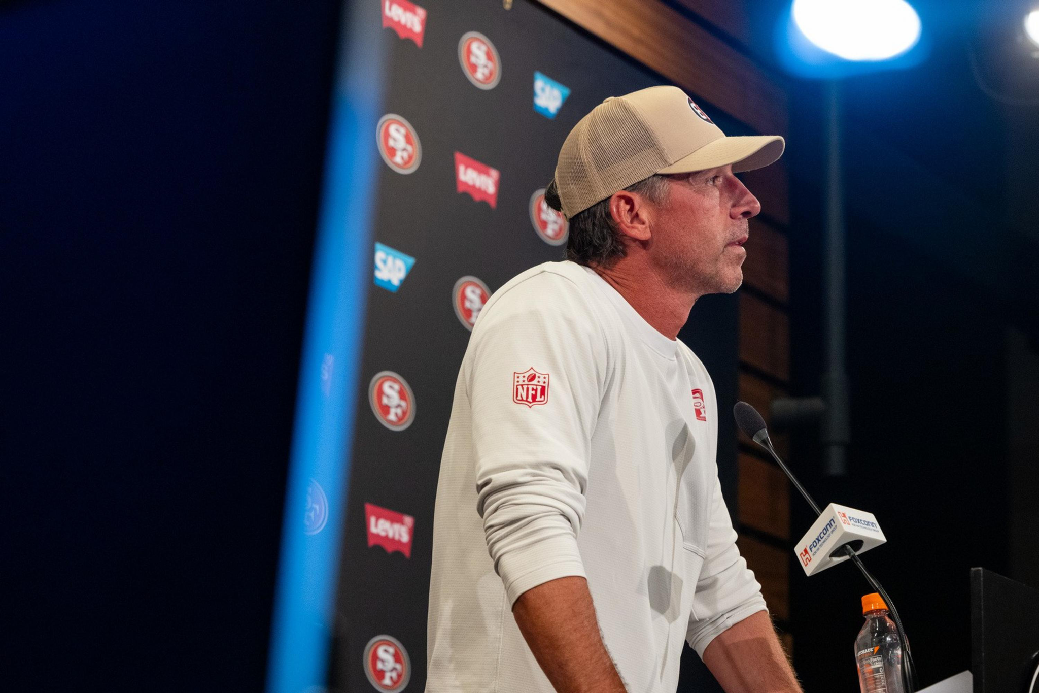 A man wearing a white long-sleeve shirt and beige cap speaks at a San Francisco 49ers press conference podium with a water bottle nearby.