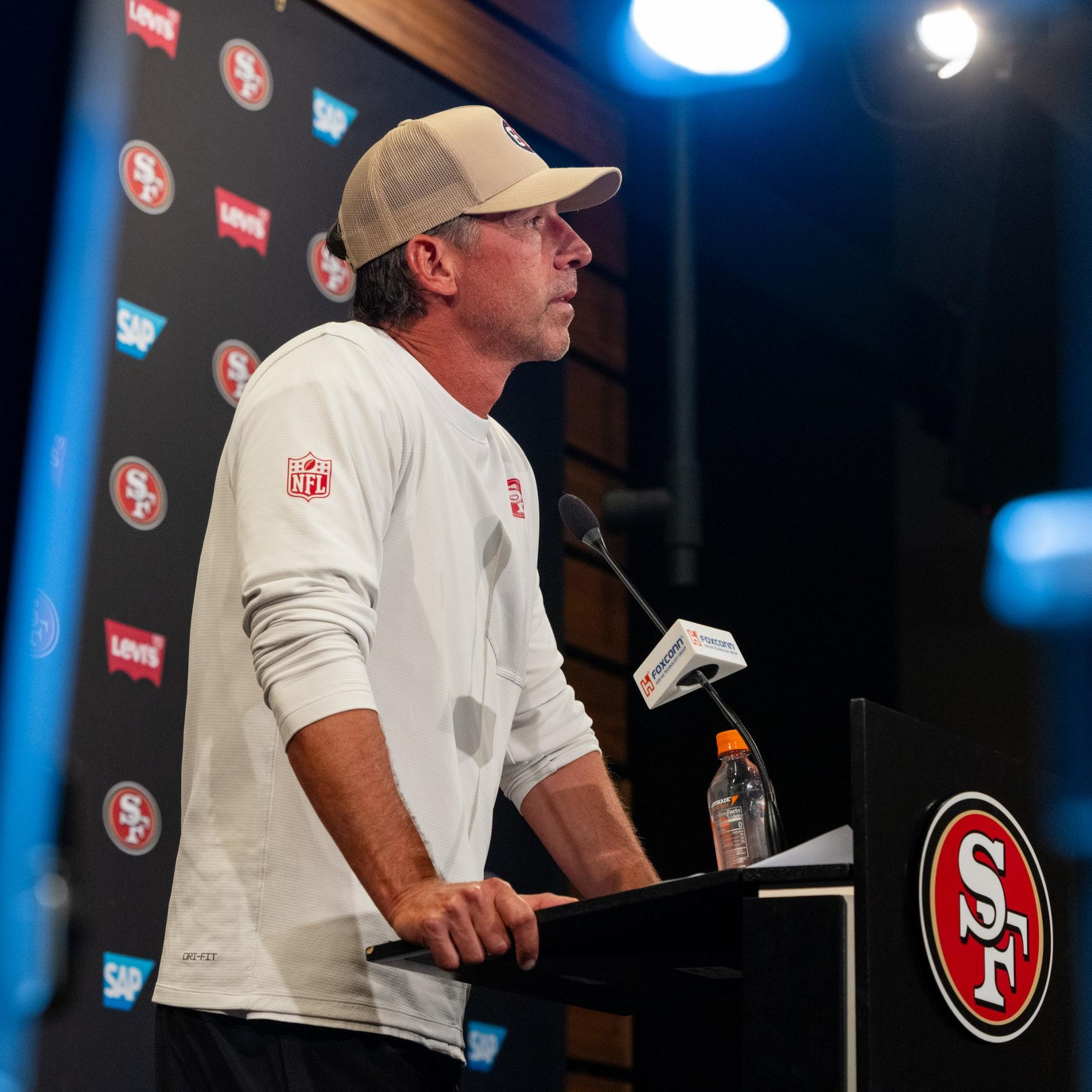 A man wearing a white long-sleeve shirt and beige cap speaks at a San Francisco 49ers press conference podium with a water bottle nearby.