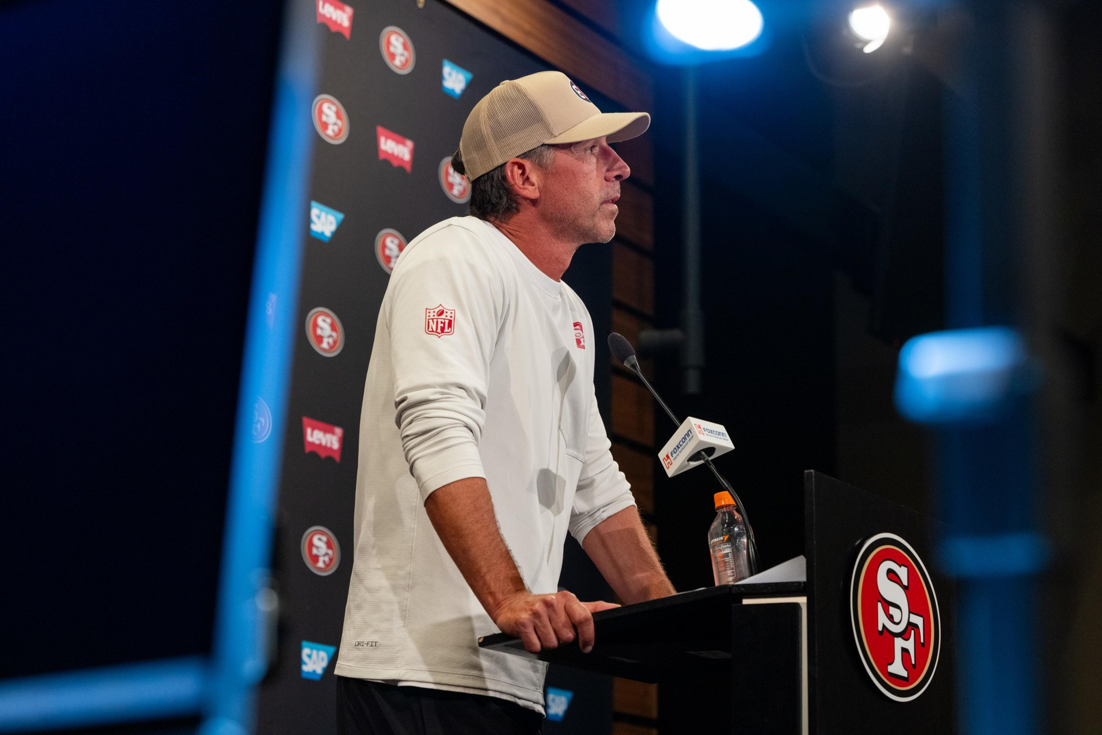 A man wearing a white long-sleeve shirt and beige cap speaks at a San Francisco 49ers press conference podium with a water bottle nearby.