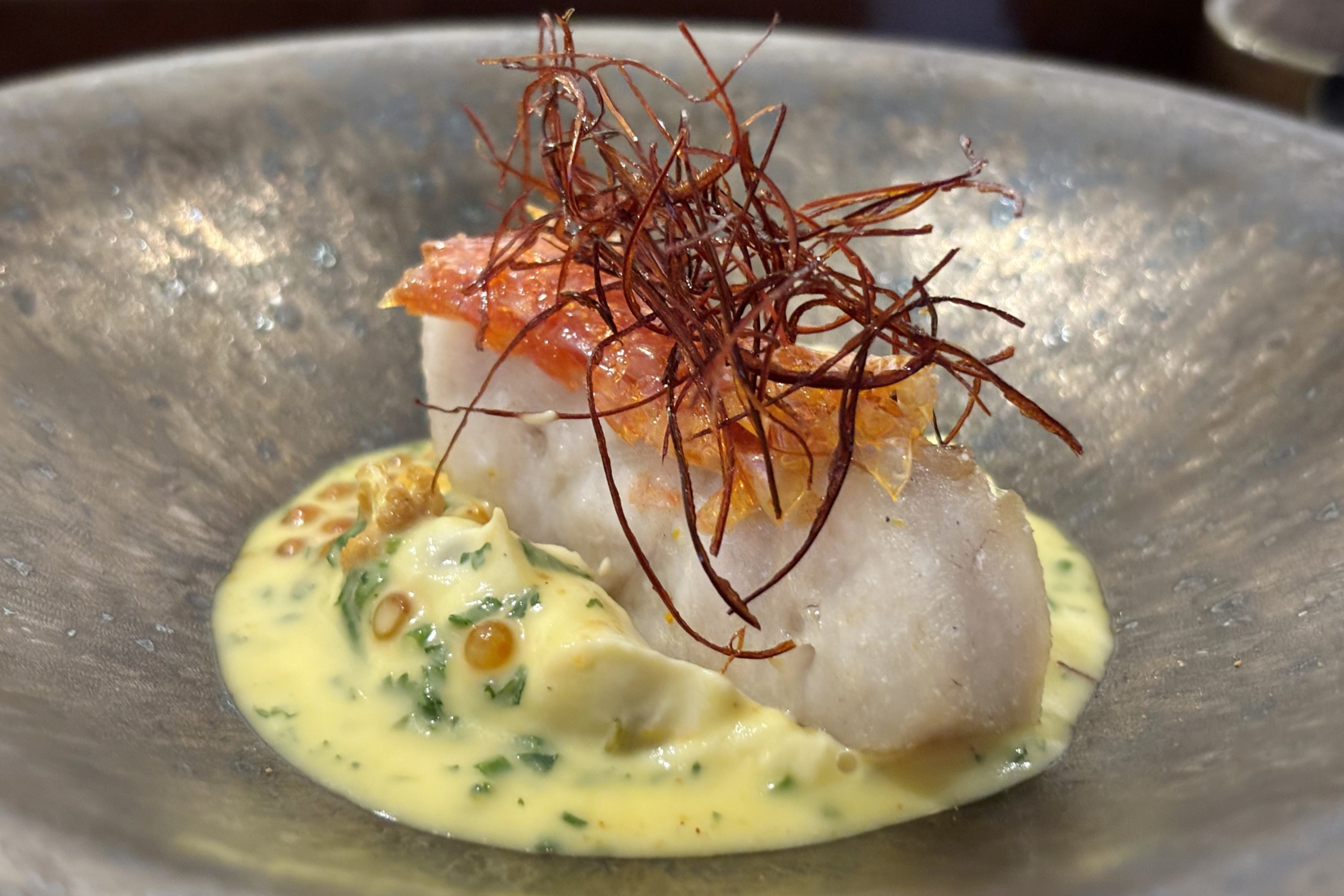 A piece of fish is served in a bowl atop a creamy, herb-speckled yellow sauce, garnished with red, crispy strands and orange fish eggs.
