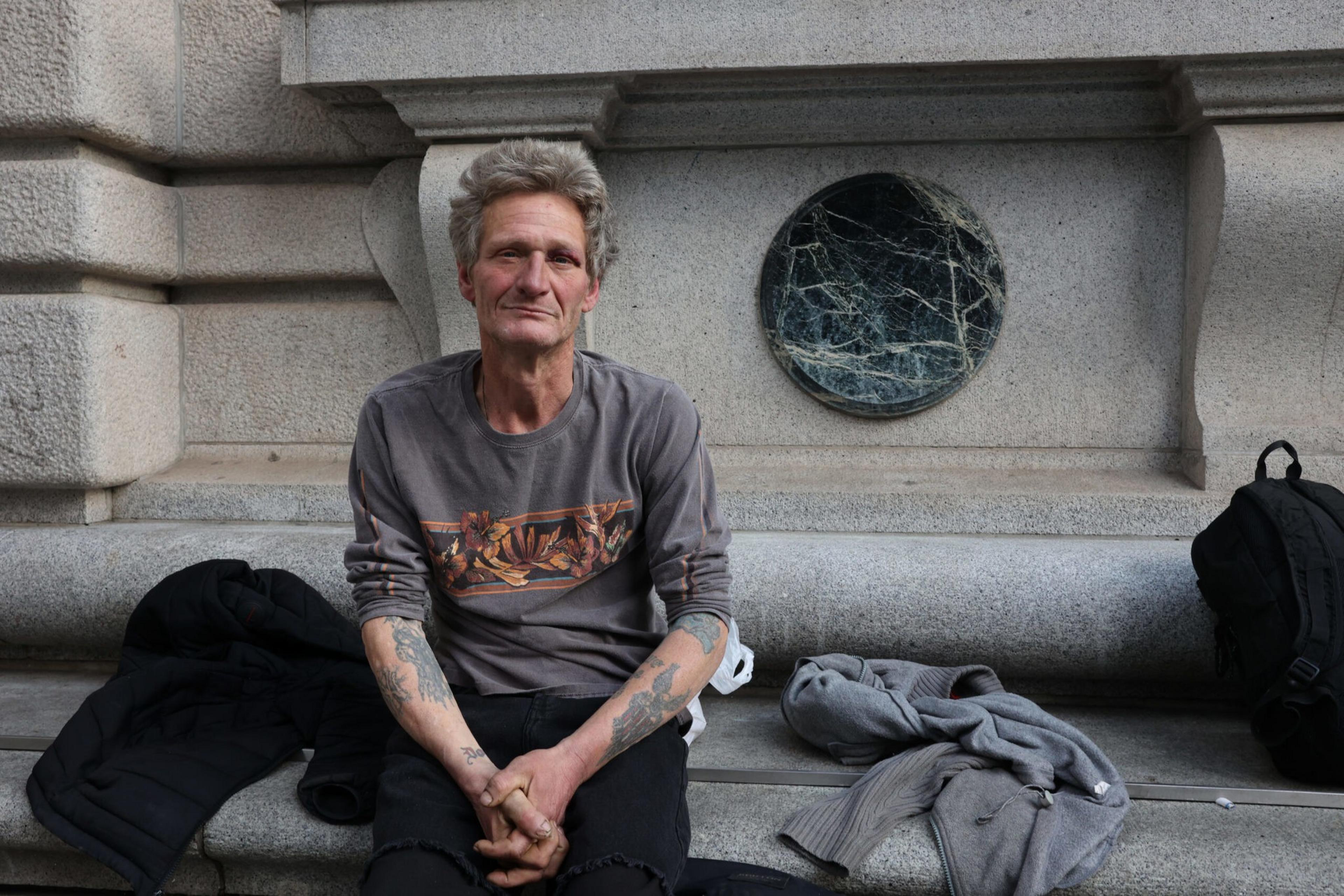 A man with gray hair and tattoos on his forearms sits on a stone bench, wearing a gray long-sleeve shirt and black pants, surrounded by clothes and backpacks.