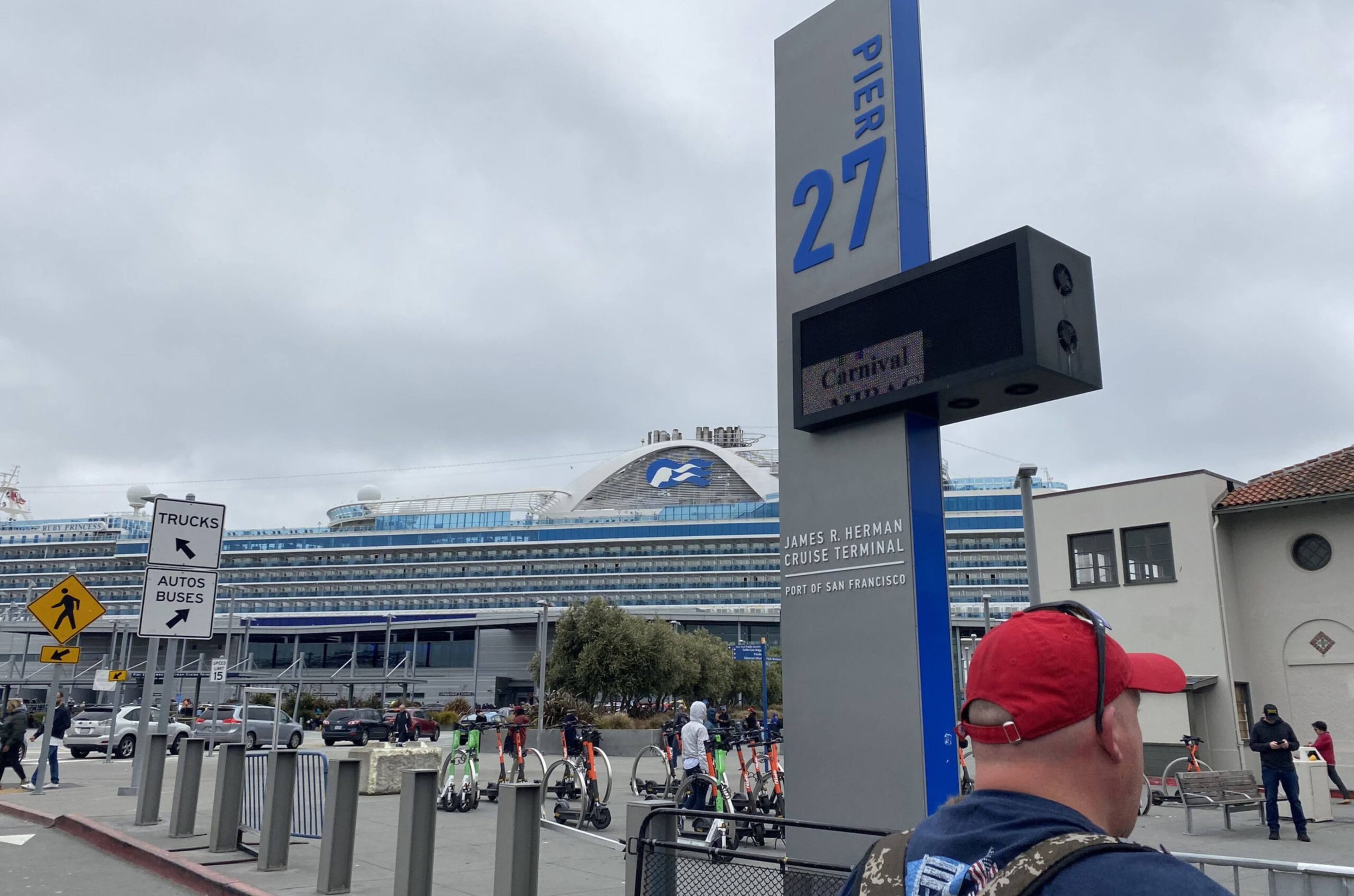 A large cruise ship docked at Pier 27, with a sign marking the James R. Herman Cruise Terminal, people walking, and electric scooters nearby.