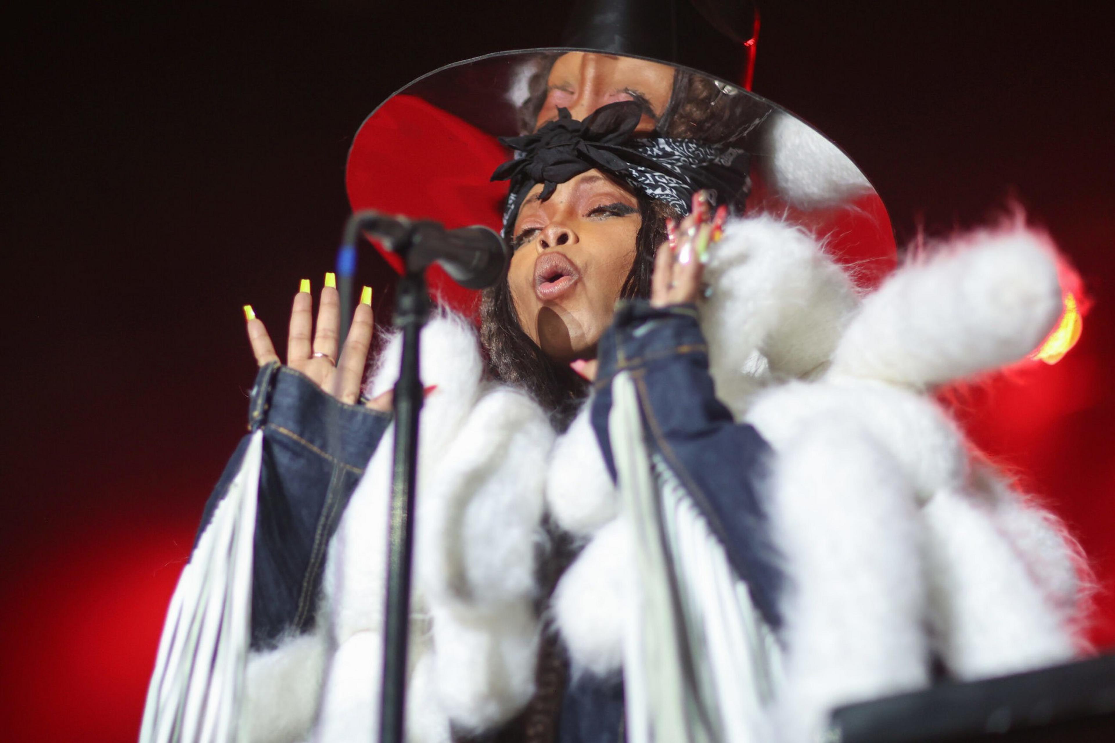 A performer with long nails, a black bandana, and a fluffy white coat sings into a microphone under red stage lighting.