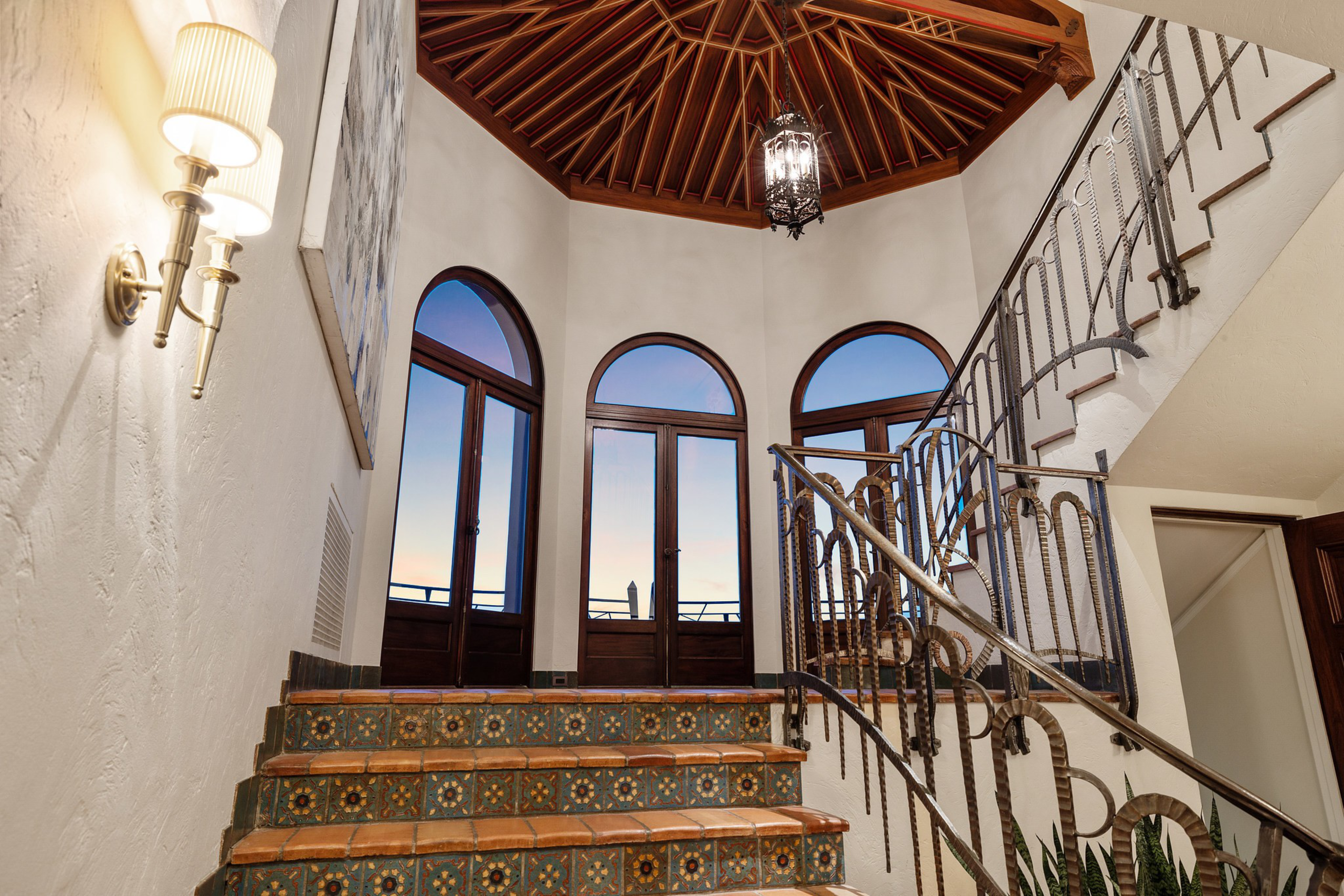 A tiled staircase leads up to three arched wooden doors with glass panels beneath a wooden vaulted ceiling and ornate hanging lantern.