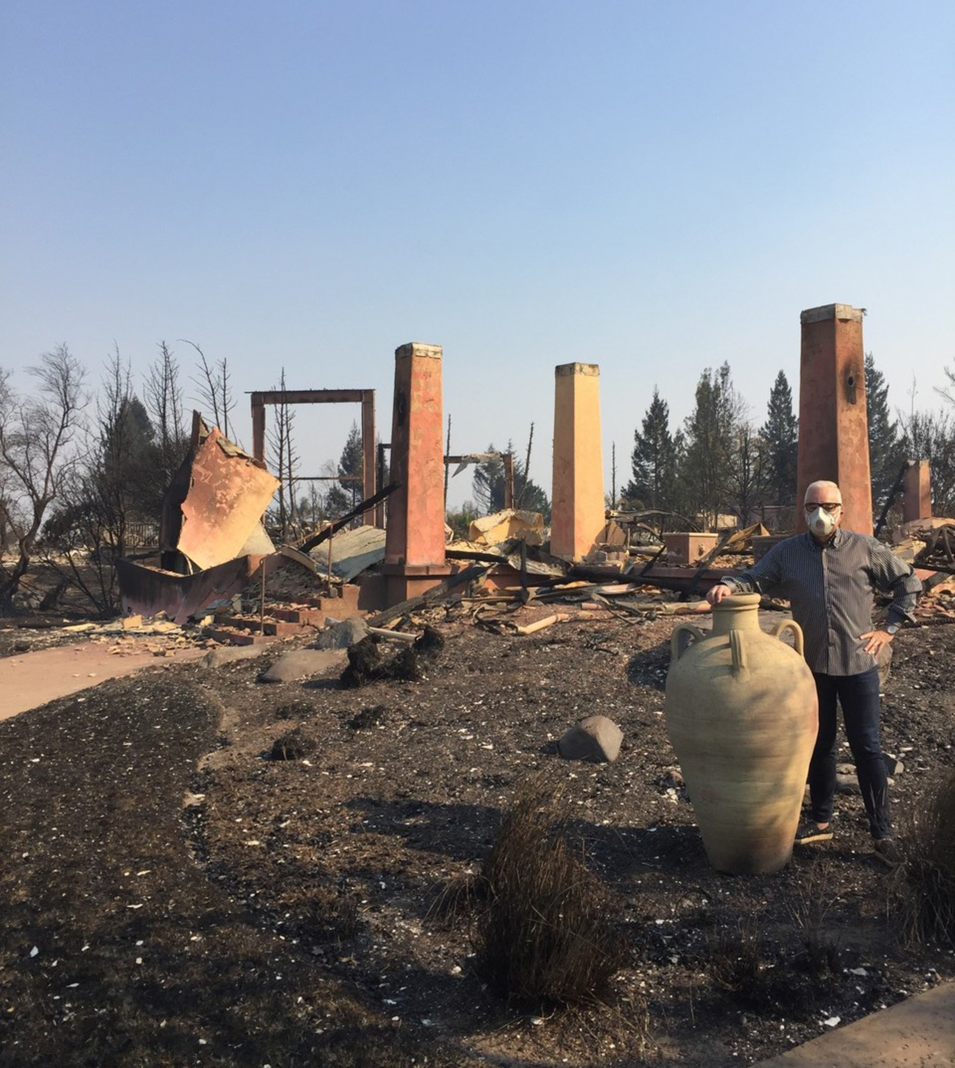 A person wearing a face mask stands next to a large ceramic pot in front of charred, collapsed remains of a building with only a few upright columns left standing.