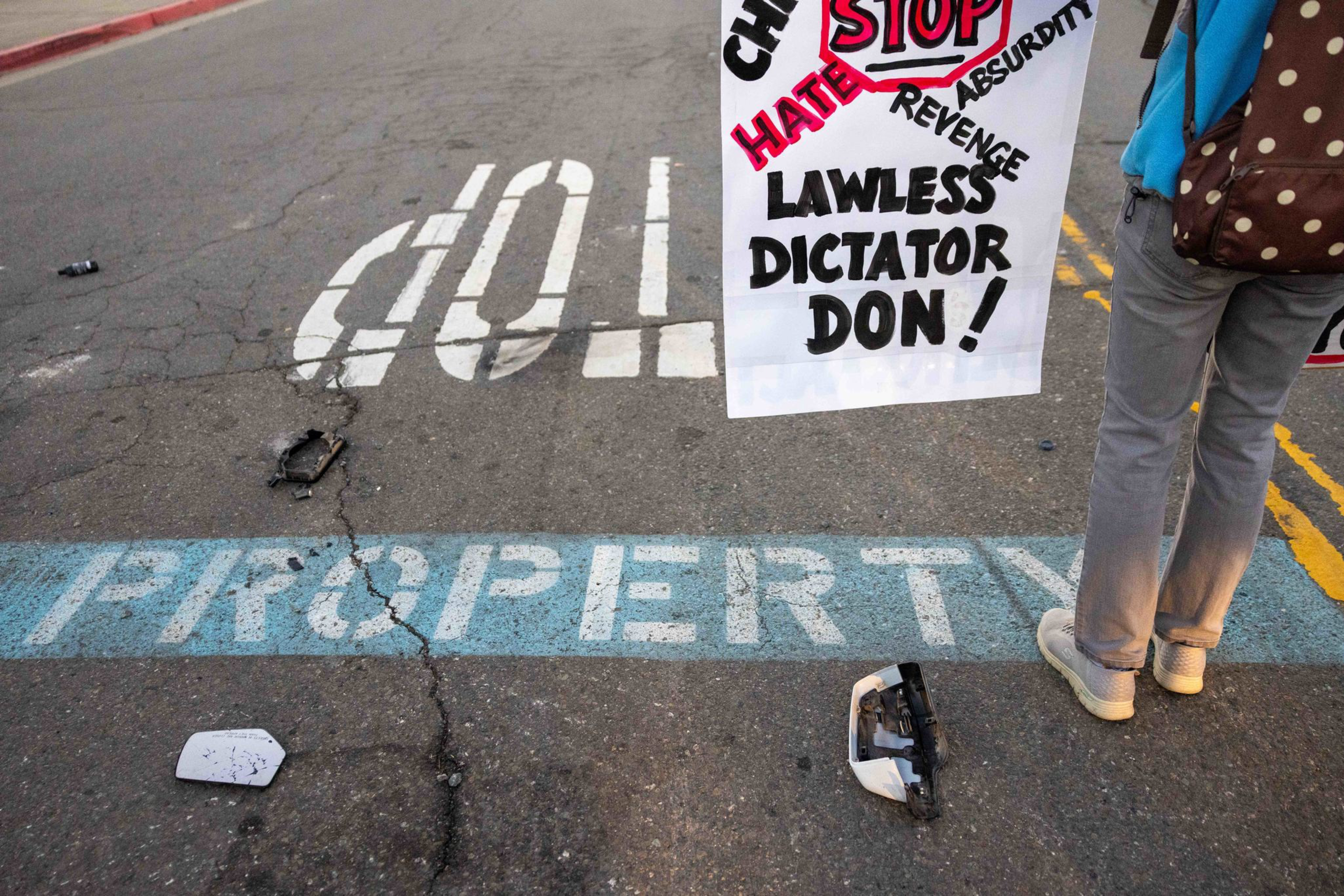 A person stands on a street near the words “STOP” and “PROPERTY” painted on the ground, holding a protest sign calling out a “lawless dictator.”