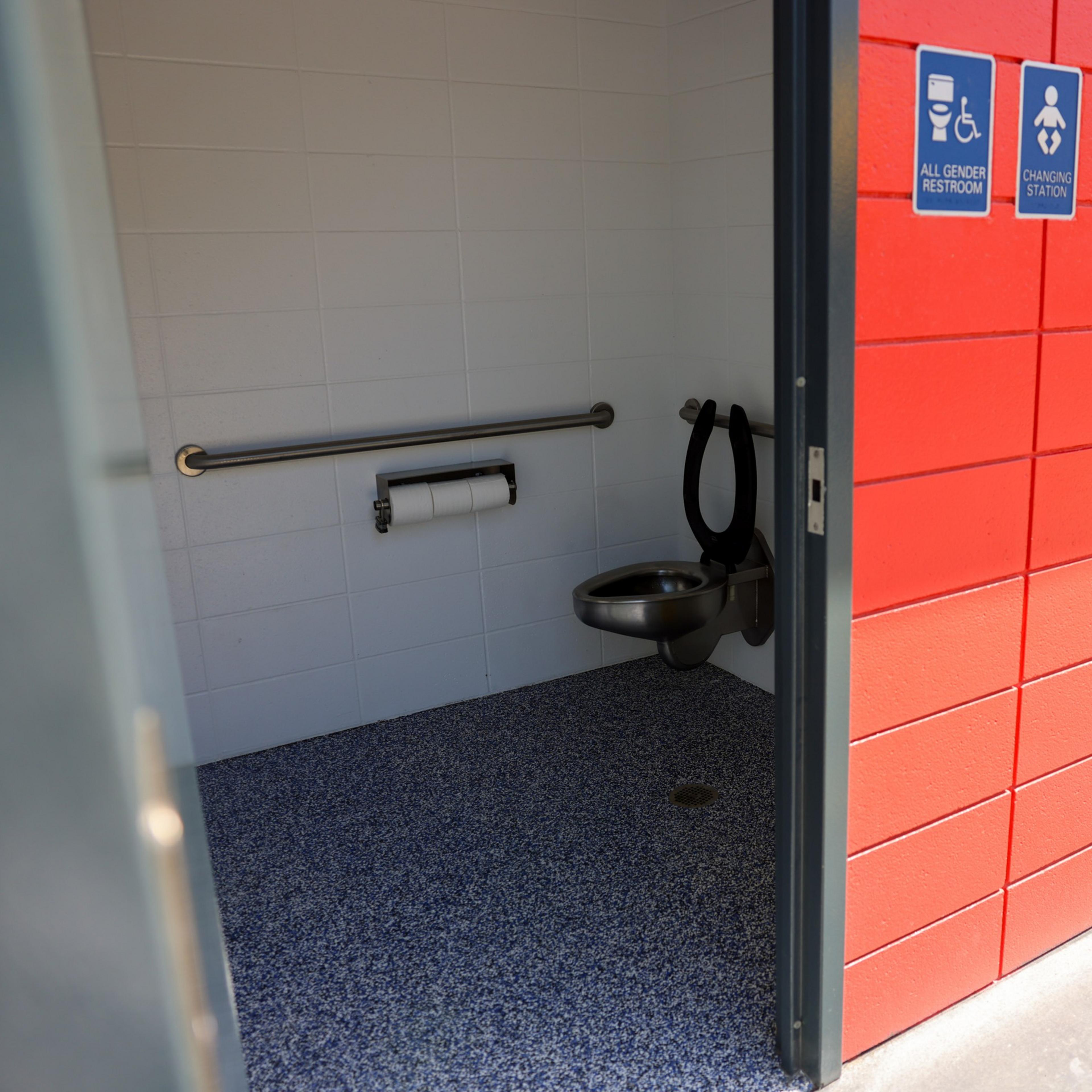An accessible restroom with white tiles, a steel toilet, handrails, and a sign indicating it's for all genders and has a changing station.