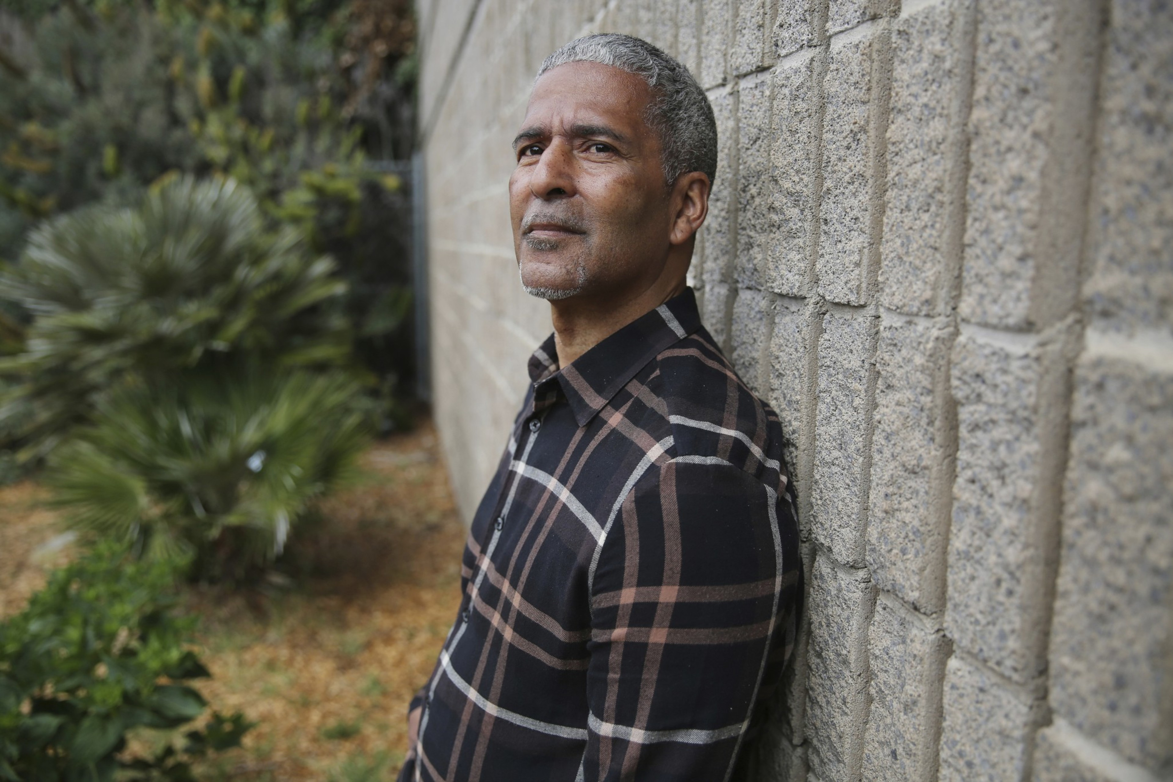 A mature man with gray hair and a goatee leans against a stone wall, wearing a black and brown plaid shirt, outdoors near greenery.