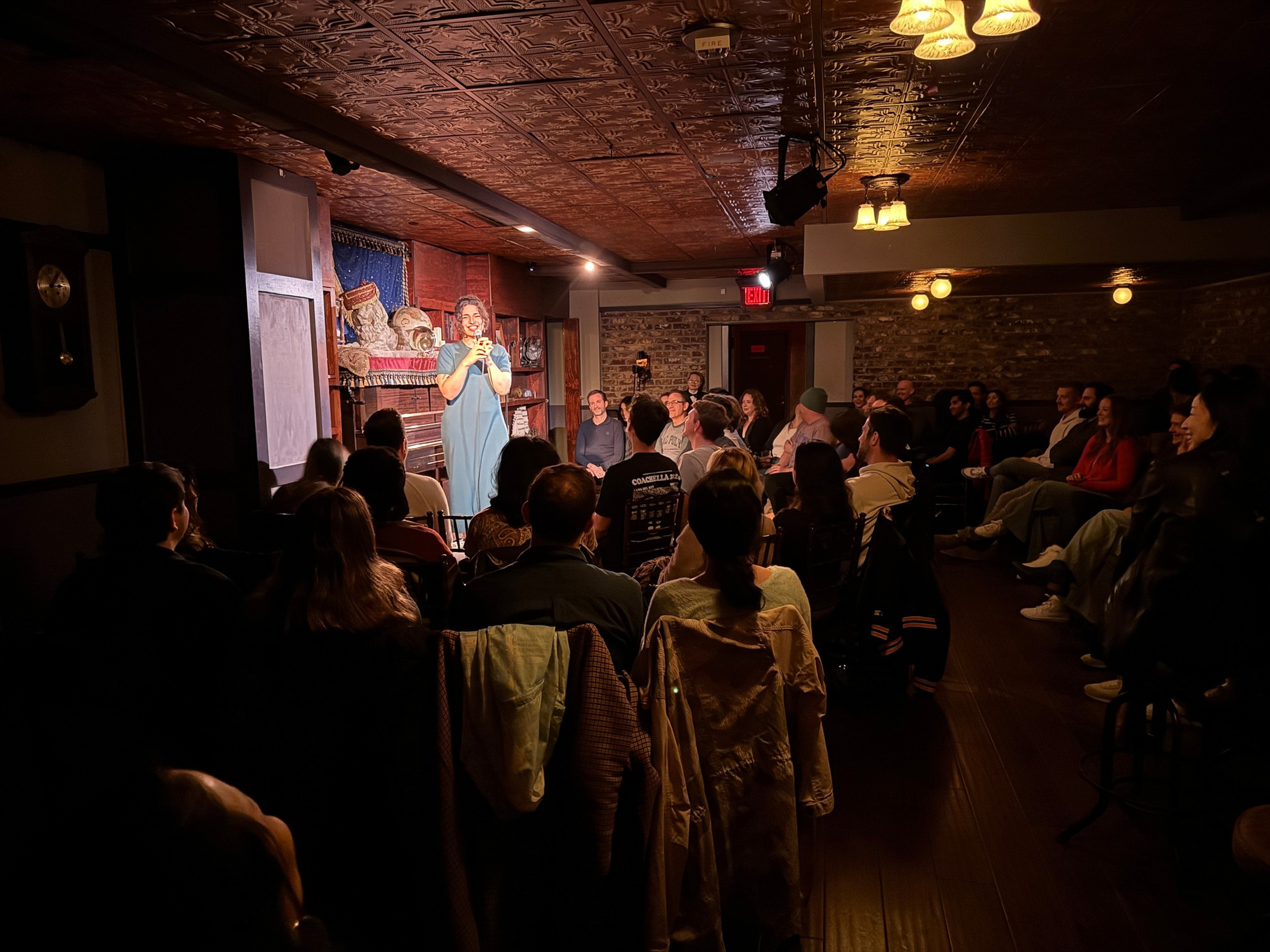 A woman in a blue dress stands on a small stage performing comedy to a seated audience in a cozy, dimly lit room with brick walls and vintage decor.