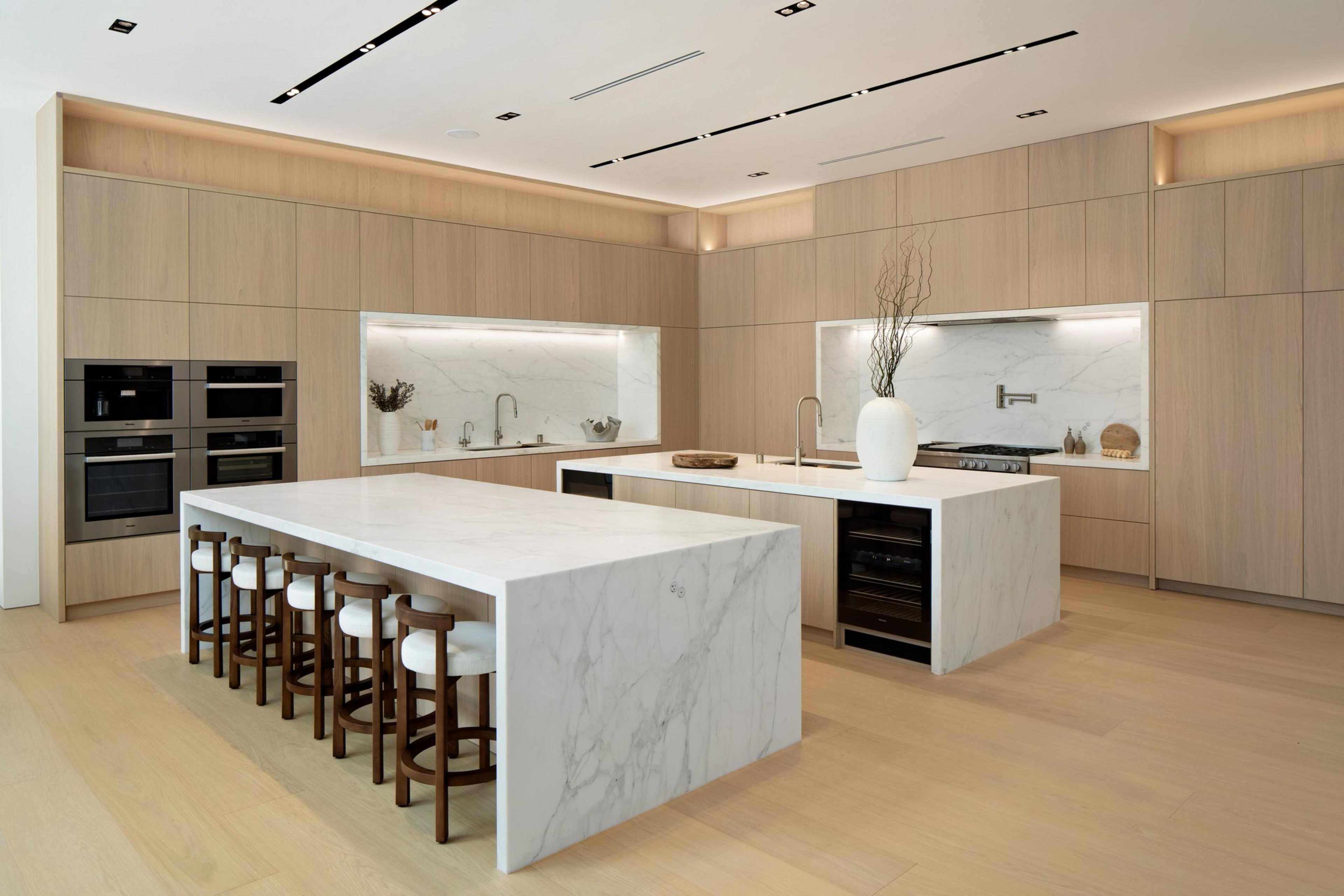 A modern kitchen features light wood cabinets, two large white marble islands, bar stools, built-in ovens, and minimalist decor with a vase on one island.