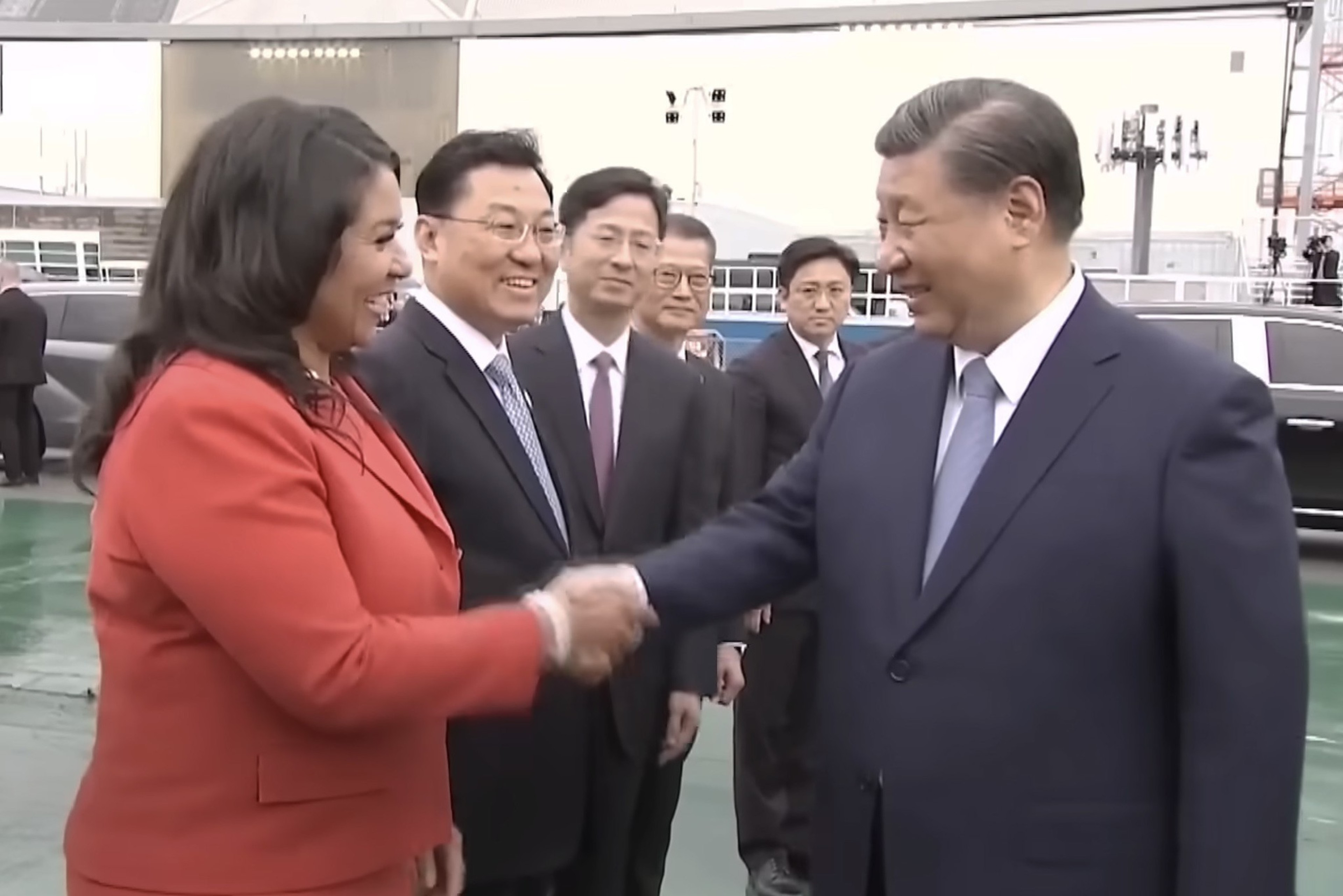 A woman in a red blazer shakes hands with a man in a dark blue suit, both smiling, with onlookers in suits behind them.