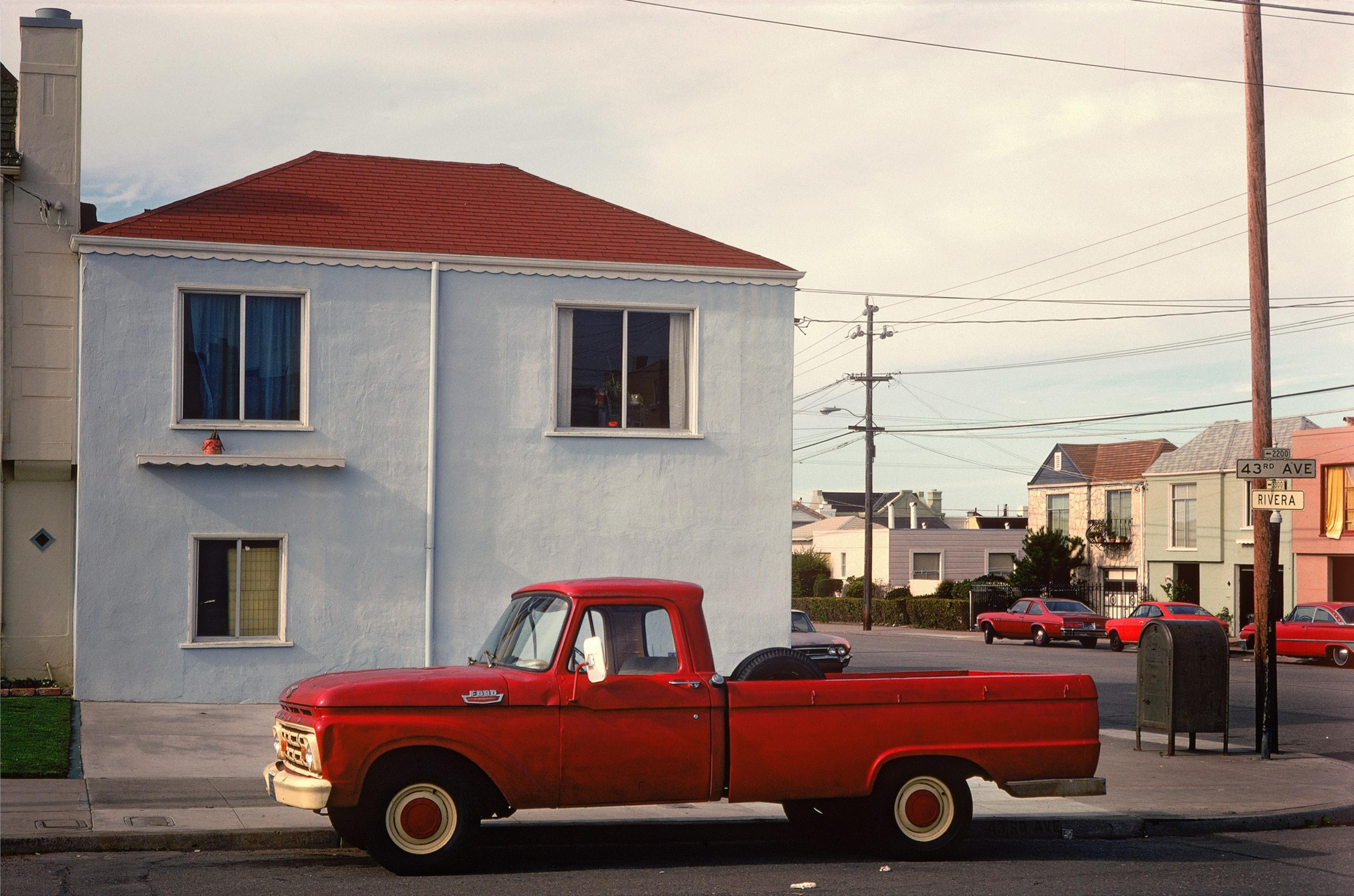 A vintage red pickup truck is parked in front of a pale blue house with a red roof, near a street corner with a sign reading &quot;43rd Ave&quot; and &quot;Rivera.&quot;