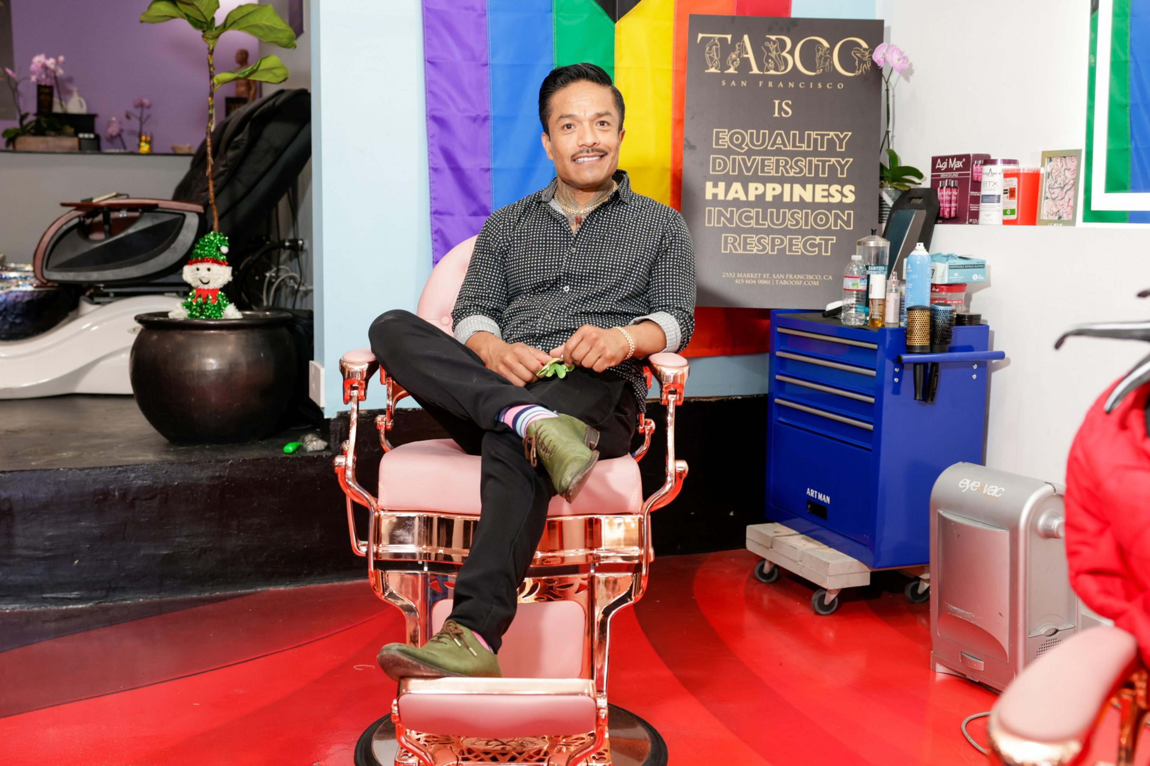 A person sits cross-legged in a barber chair, smiling. Behind them is a sign promoting equality and diversity, and a colorful flag. The environment is bright and welcoming.