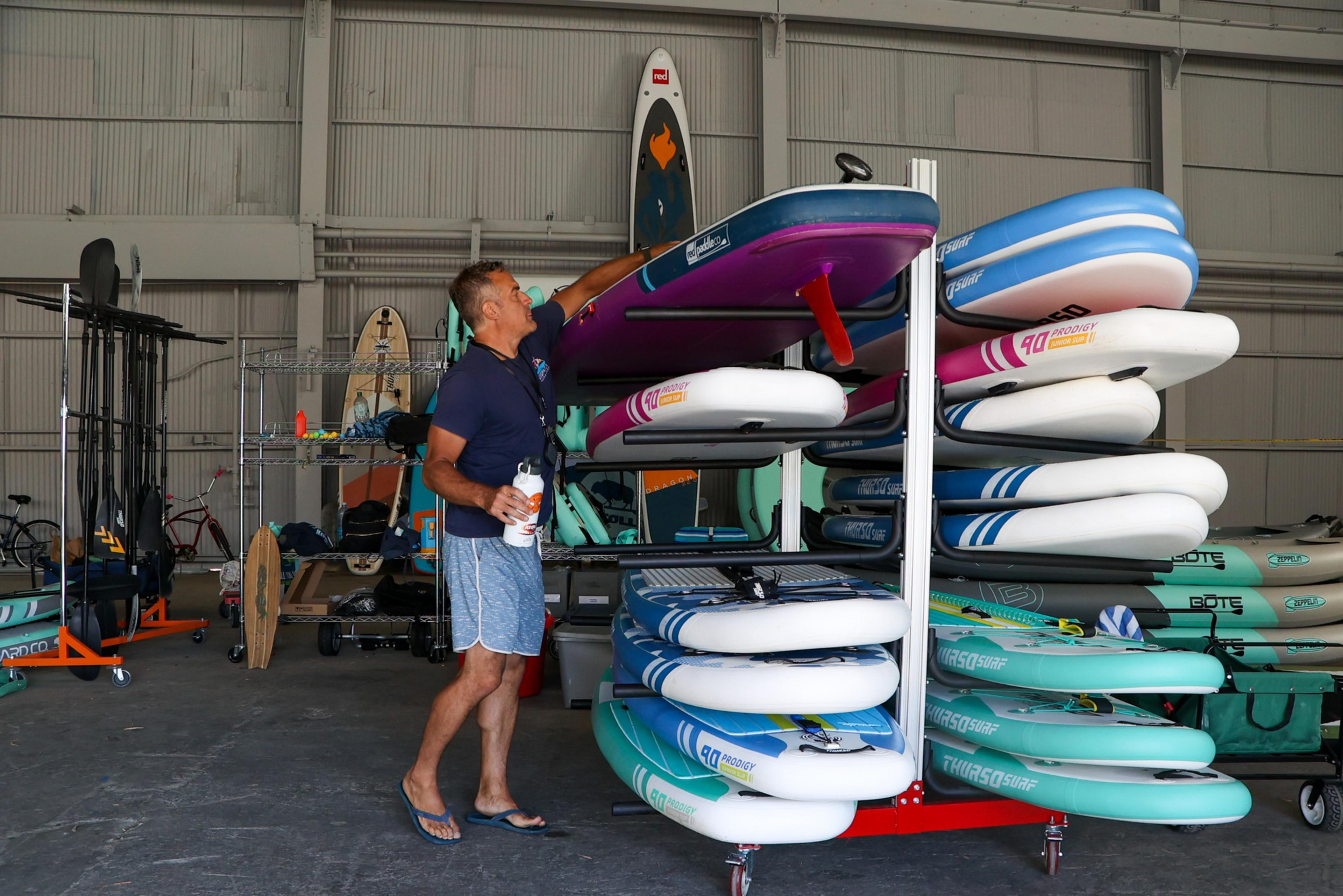 A man in a navy shirt and blue shorts arranges paddleboards stacked on a metal rack inside a storage area filled with water sports gear.