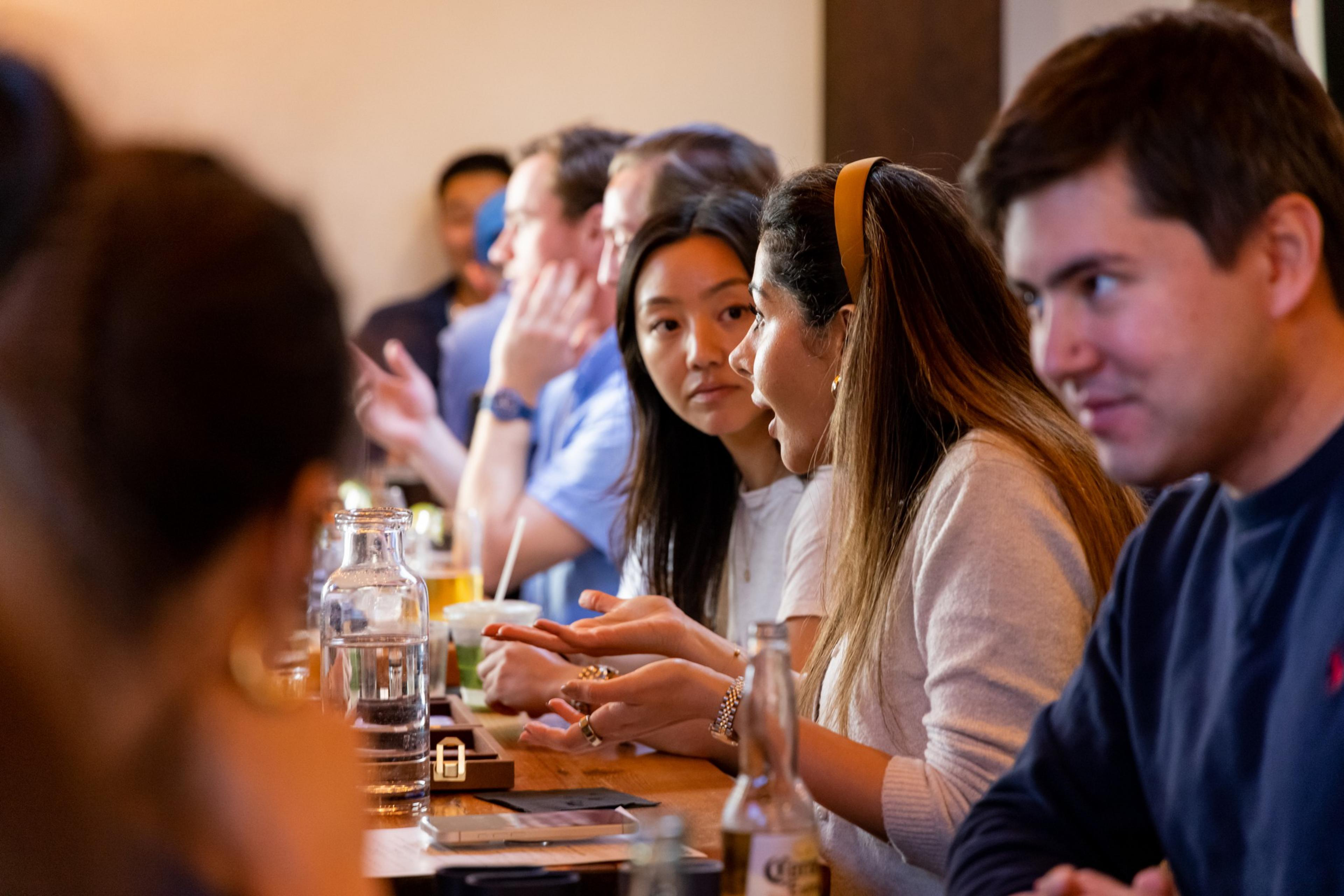 Several people sit closely at a wooden bar counter, engaged in conversation and drinking various beverages in a warm, casual setting.