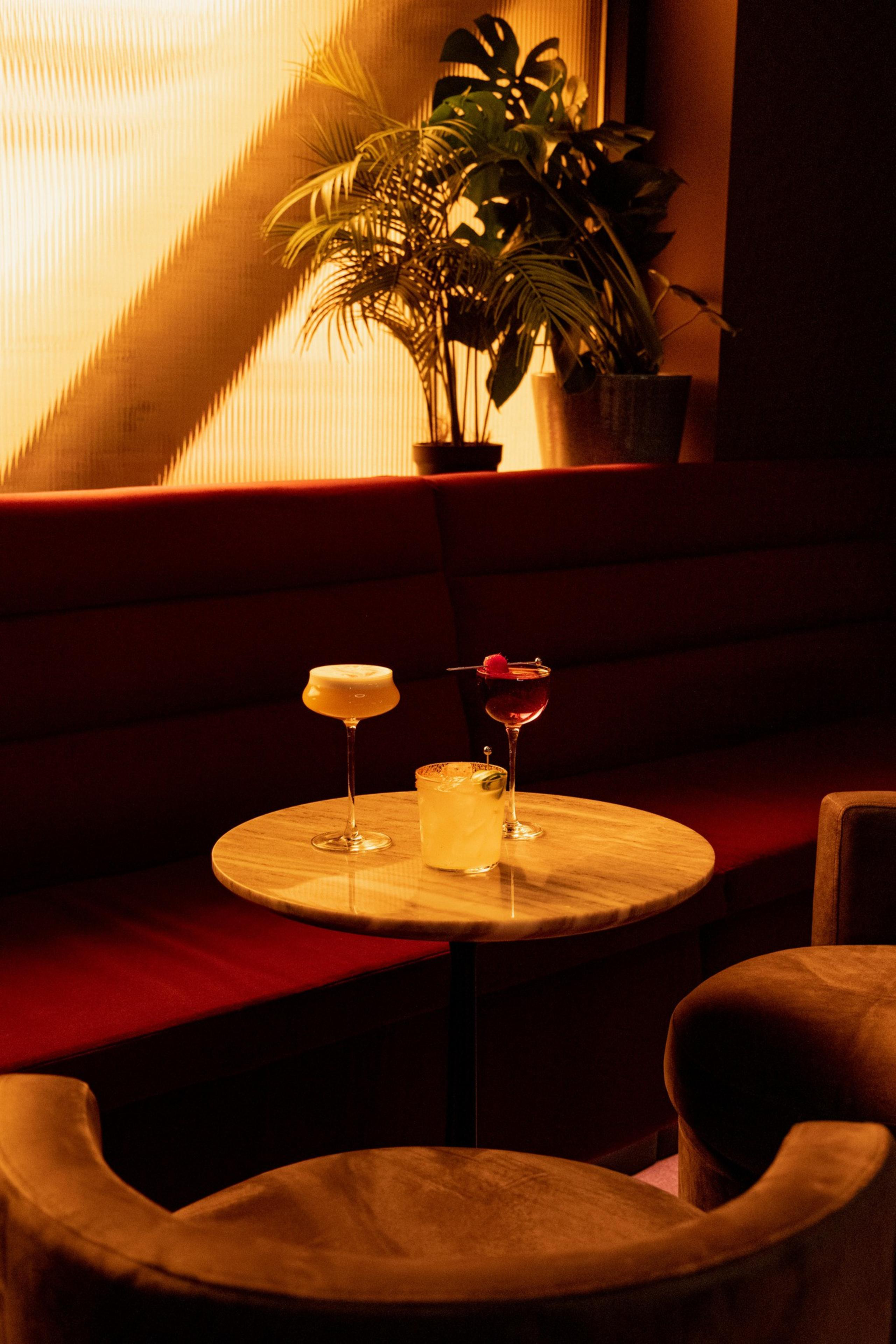 A round wooden table holds three cocktails in an intimate, warmly lit lounge with red seating and green potted plants in the background.