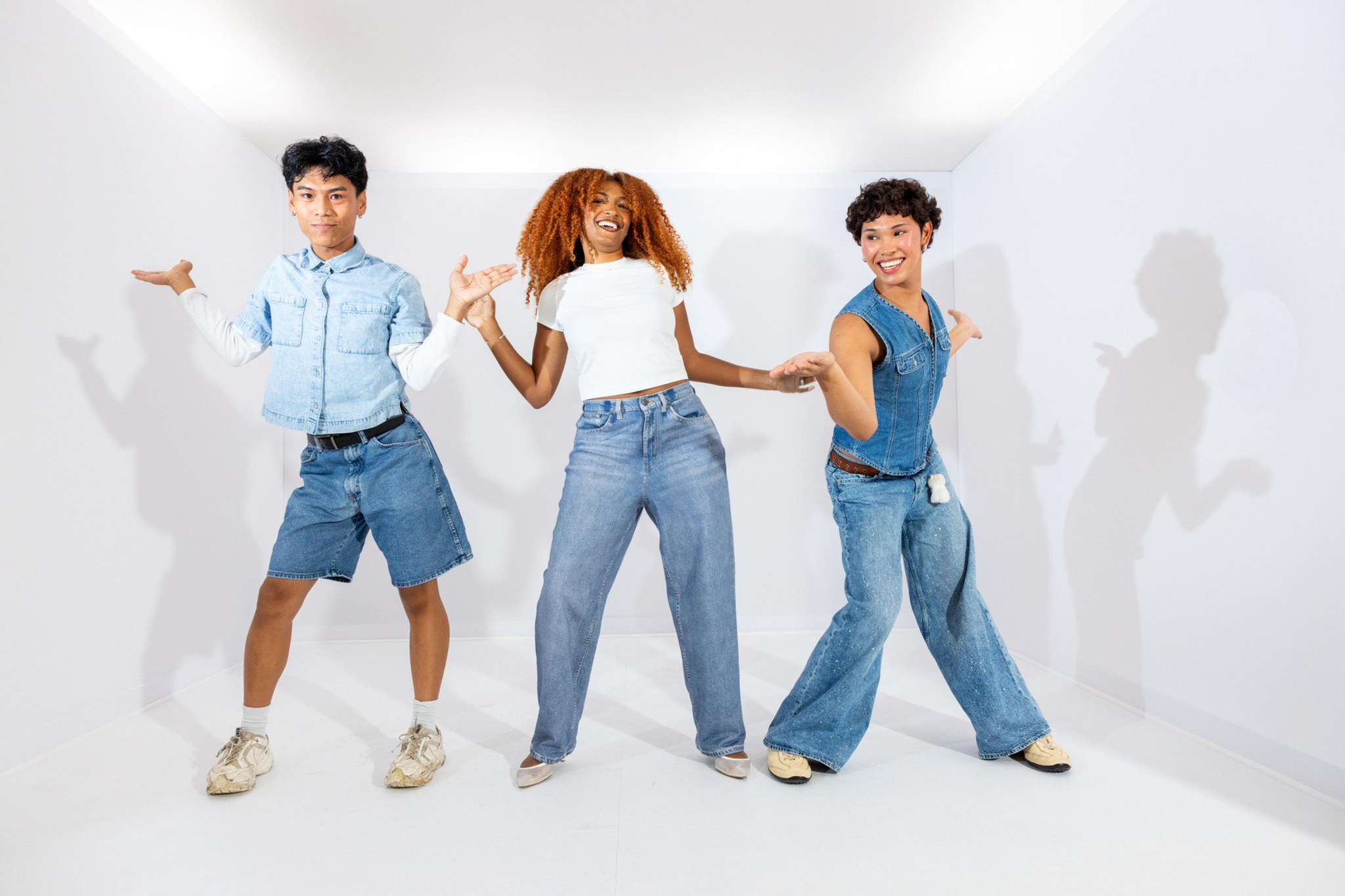 Three young people in denim outfits smile and dance in a brightly lit, empty white room with shadows cast on the walls behind them.