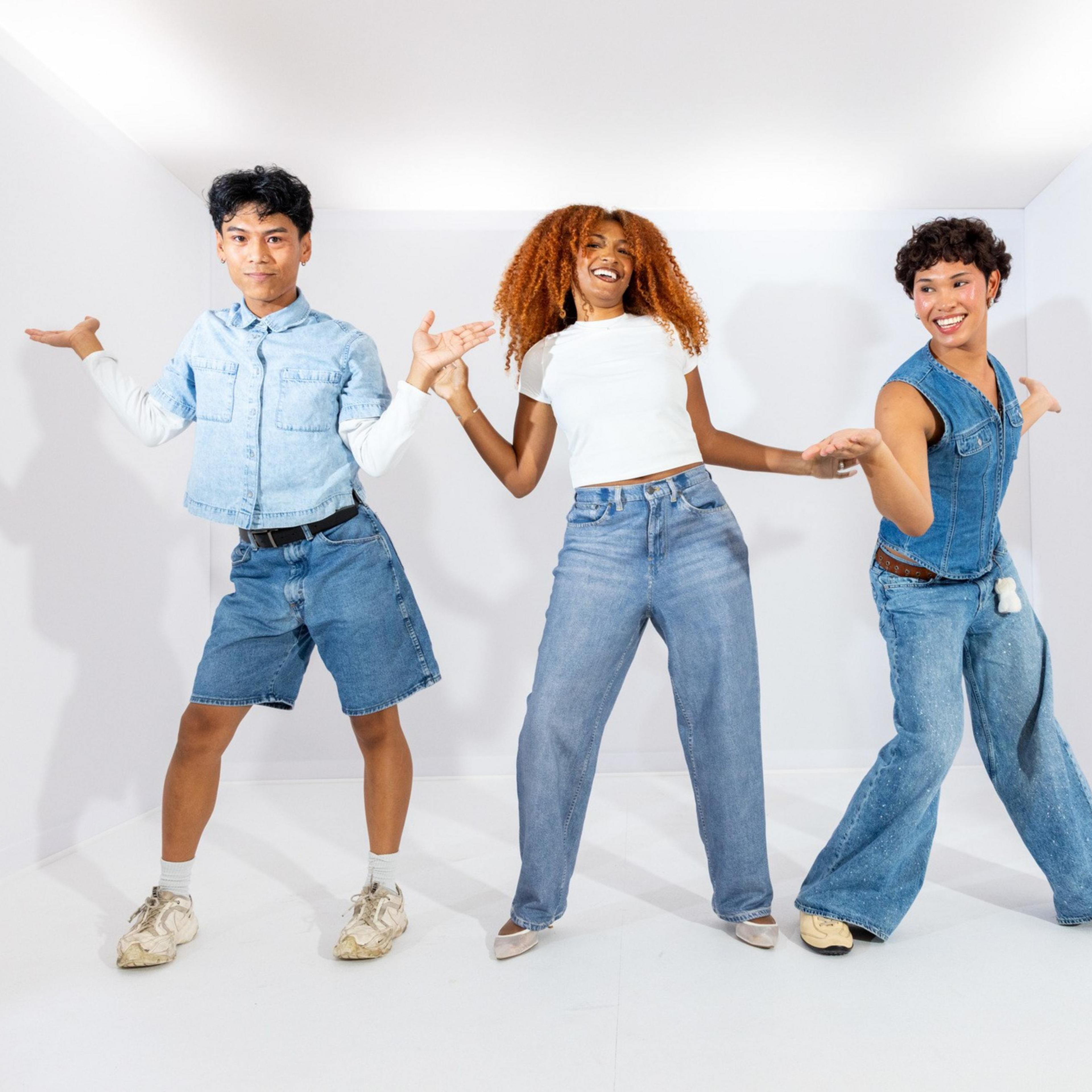 Three young people in denim outfits smile and dance in a brightly lit, empty white room with shadows cast on the walls behind them.