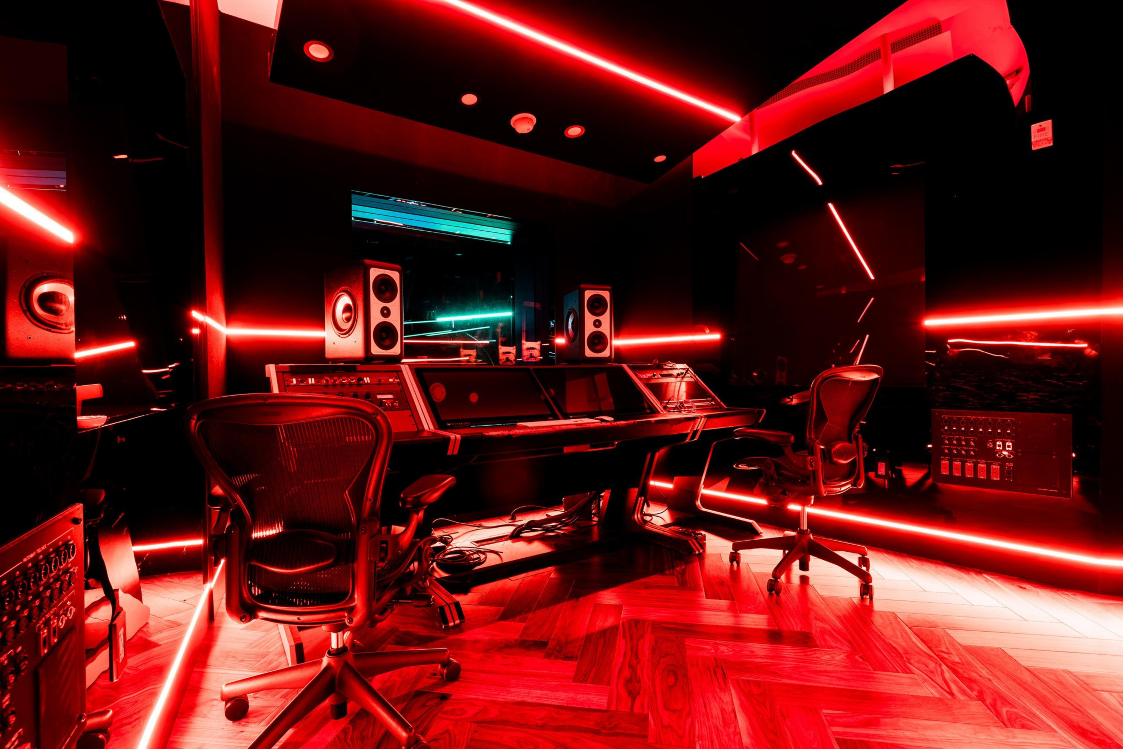 The image shows a modern recording studio with red neon lights, sleek black furniture, large speakers, a mixing console, and swivel chairs on a wooden floor.
