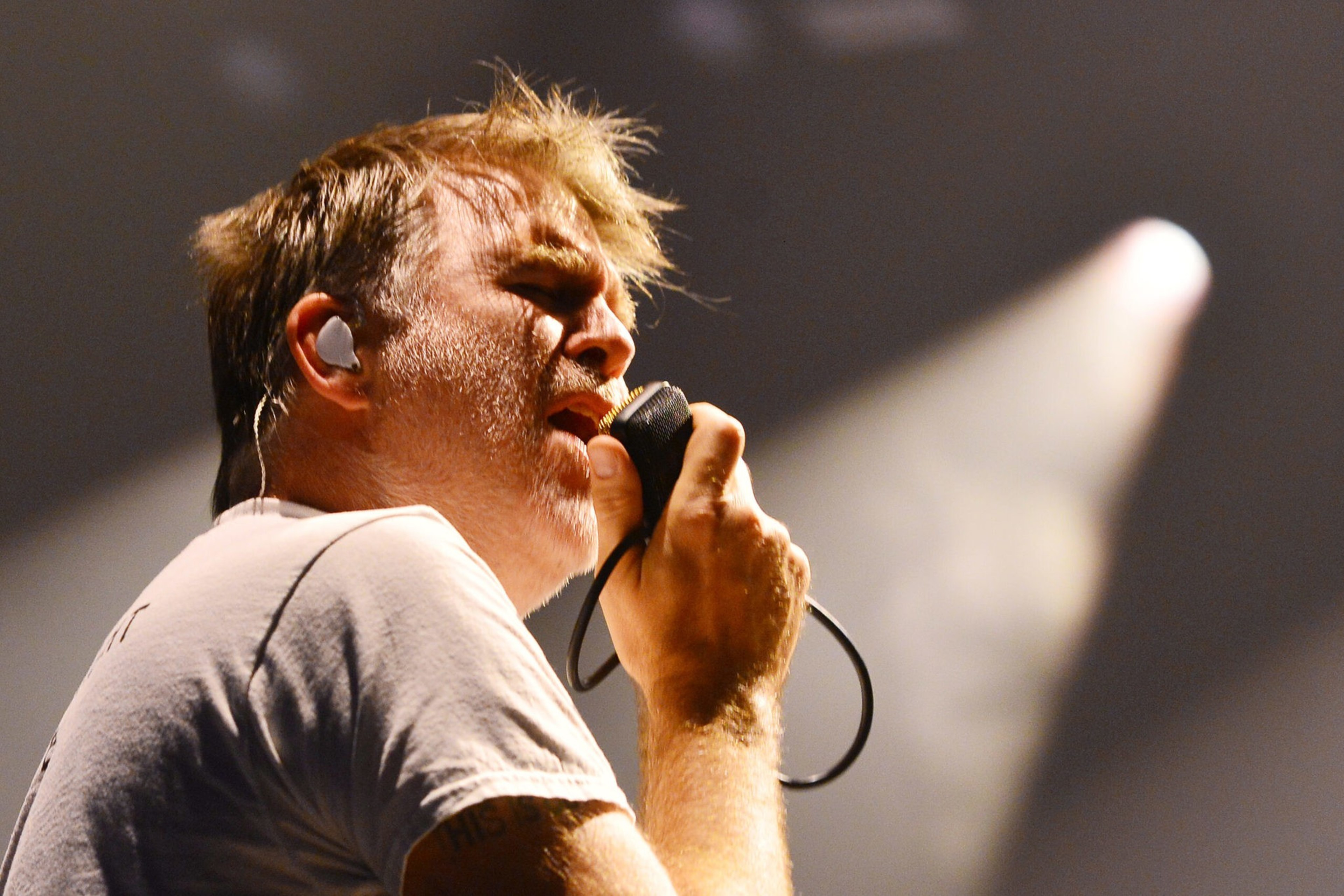 A man with blonde hair sings into a microphone with a light behind him.