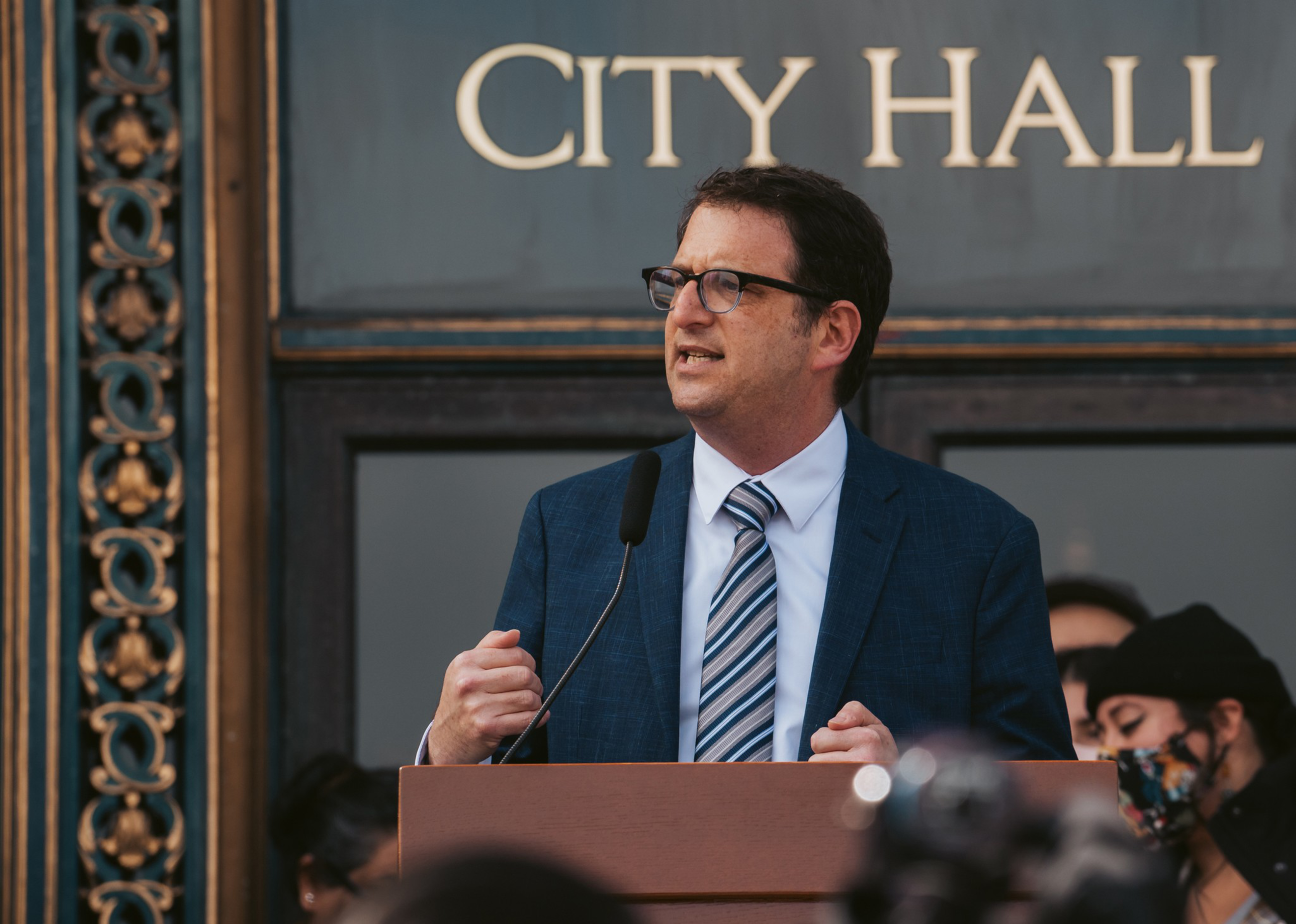 A man in a blue suit and striped tie speaks passionately at a podium outside a building labeled "City Hall."