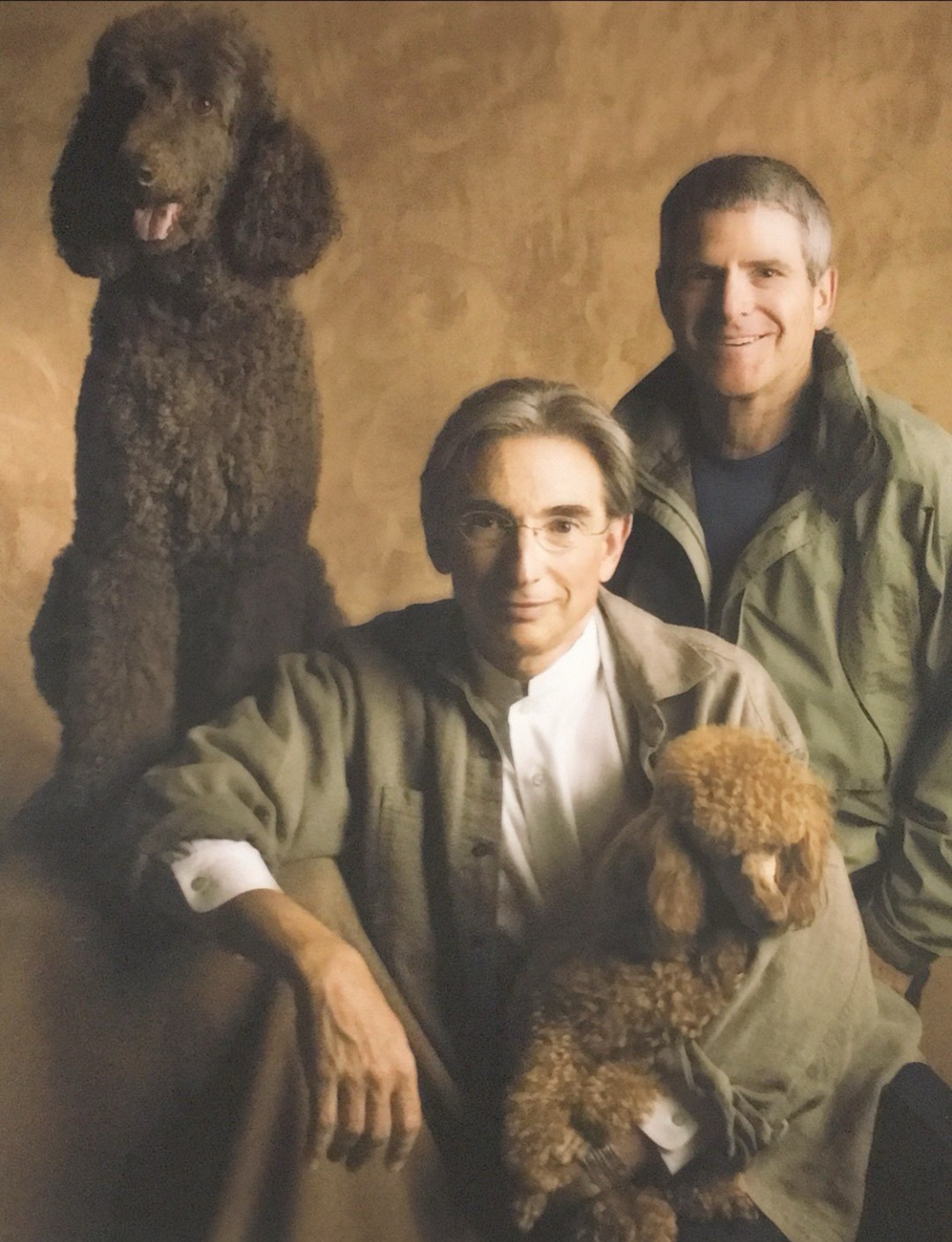 Two men sit close together; one holds a small brown poodle while a larger black poodle stands behind them against a warm, plain background.