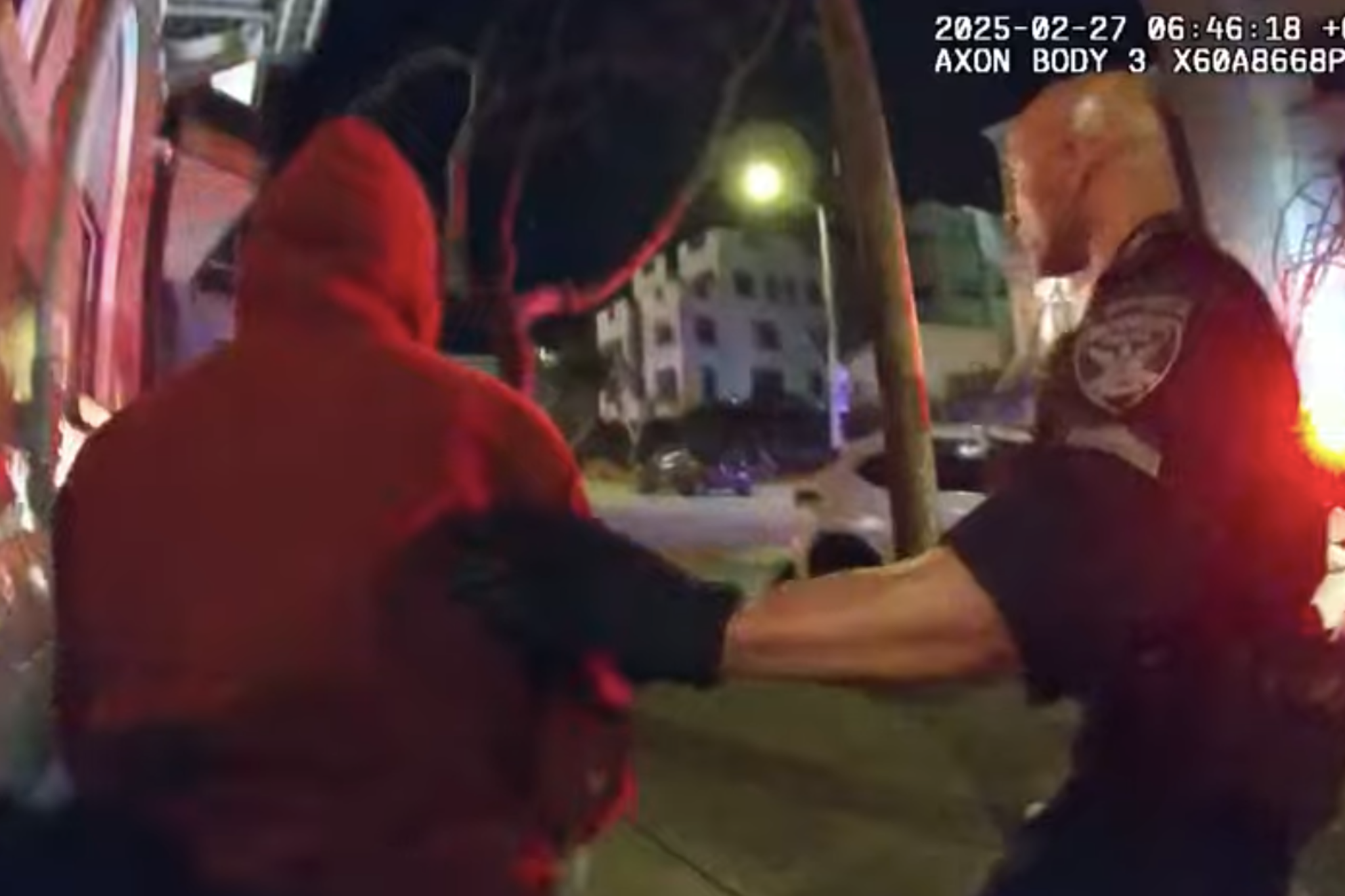 A police officer wearing black gloves holds the arm of a person in a red hoodie during a nighttime street interaction.