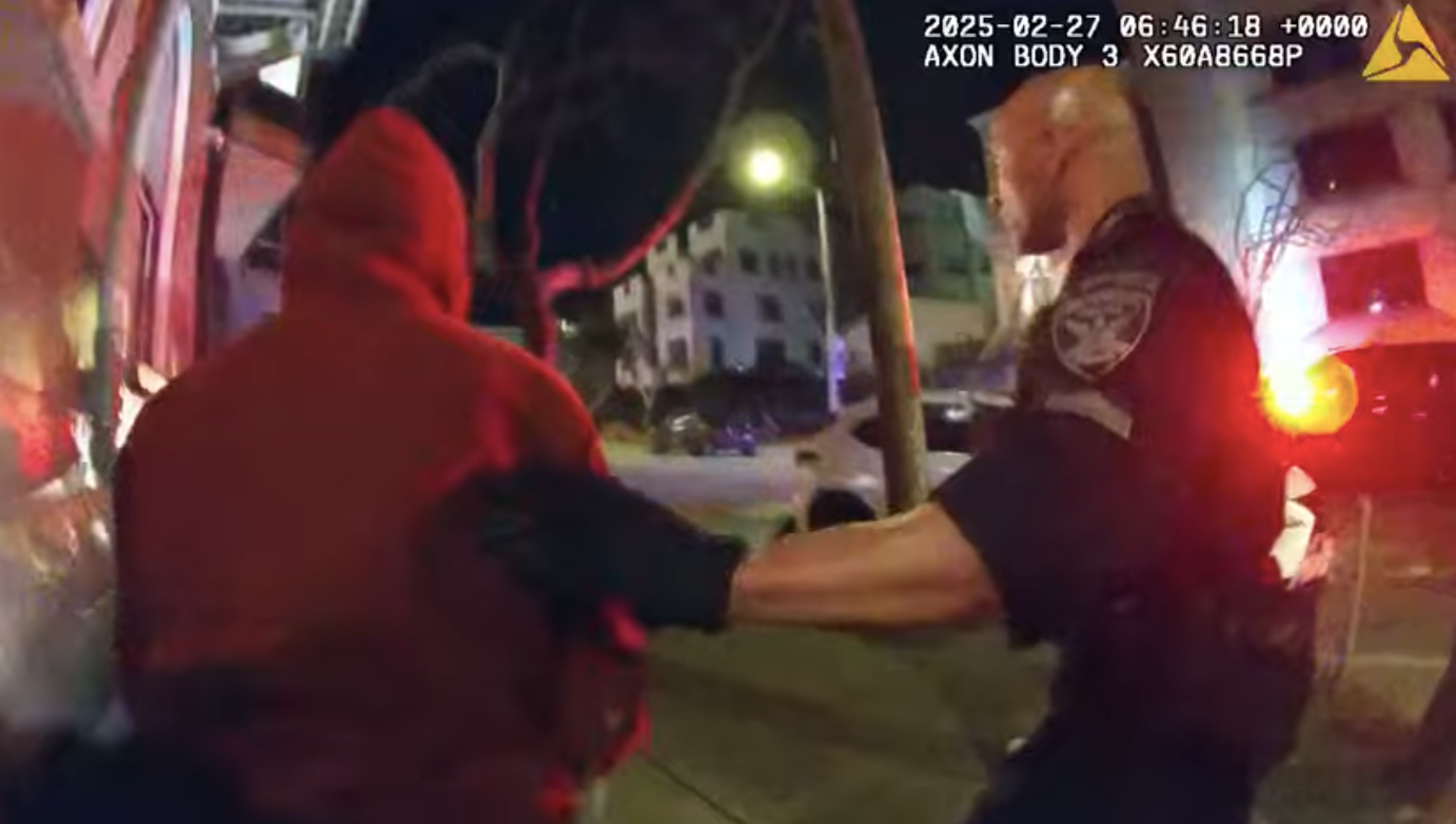 A police officer wearing black gloves holds the arm of a person in a red hoodie during a nighttime street interaction.