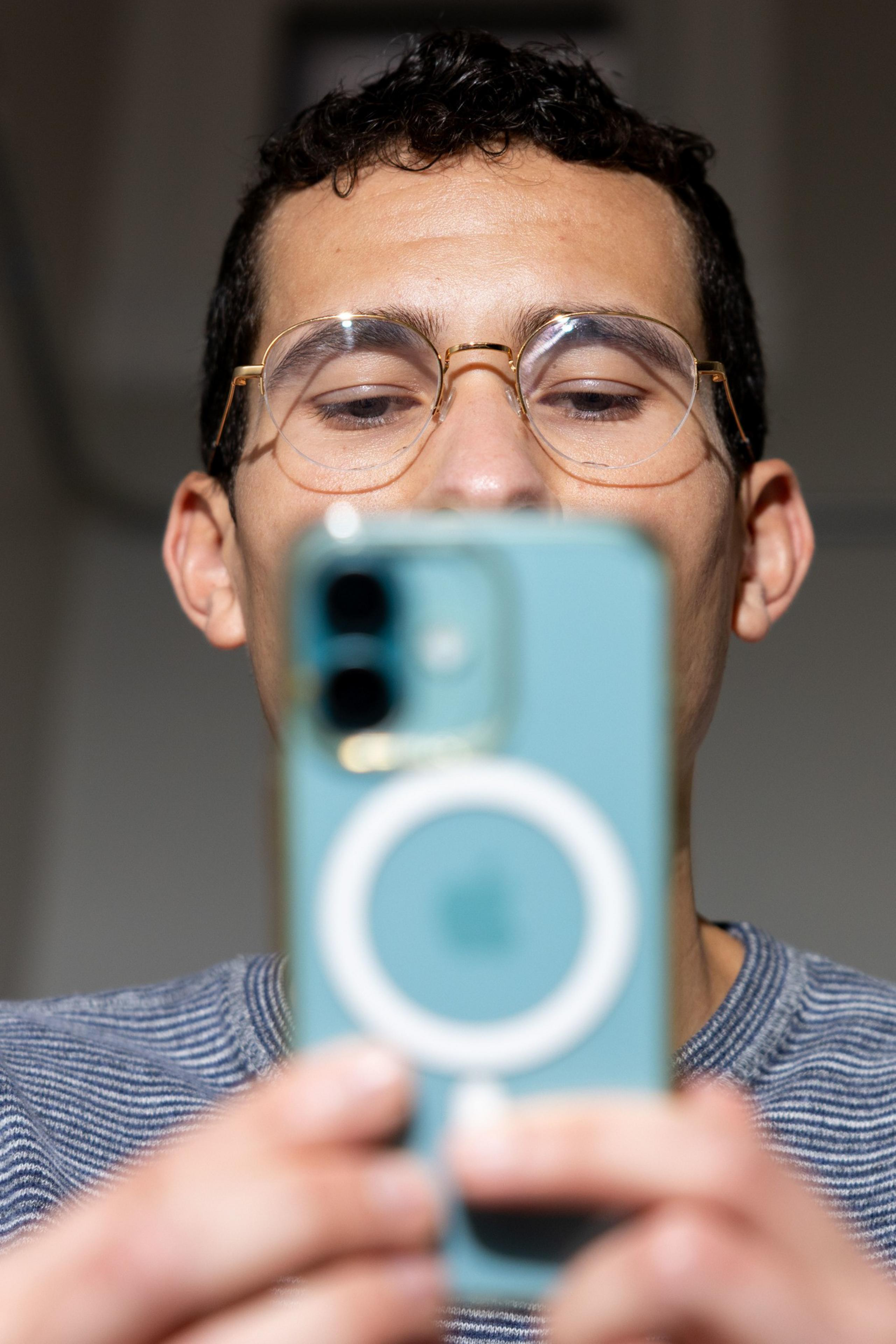 A person wearing glasses is taking a selfie with a turquoise iPhone. The focus is on the phone, with their face visible above it.