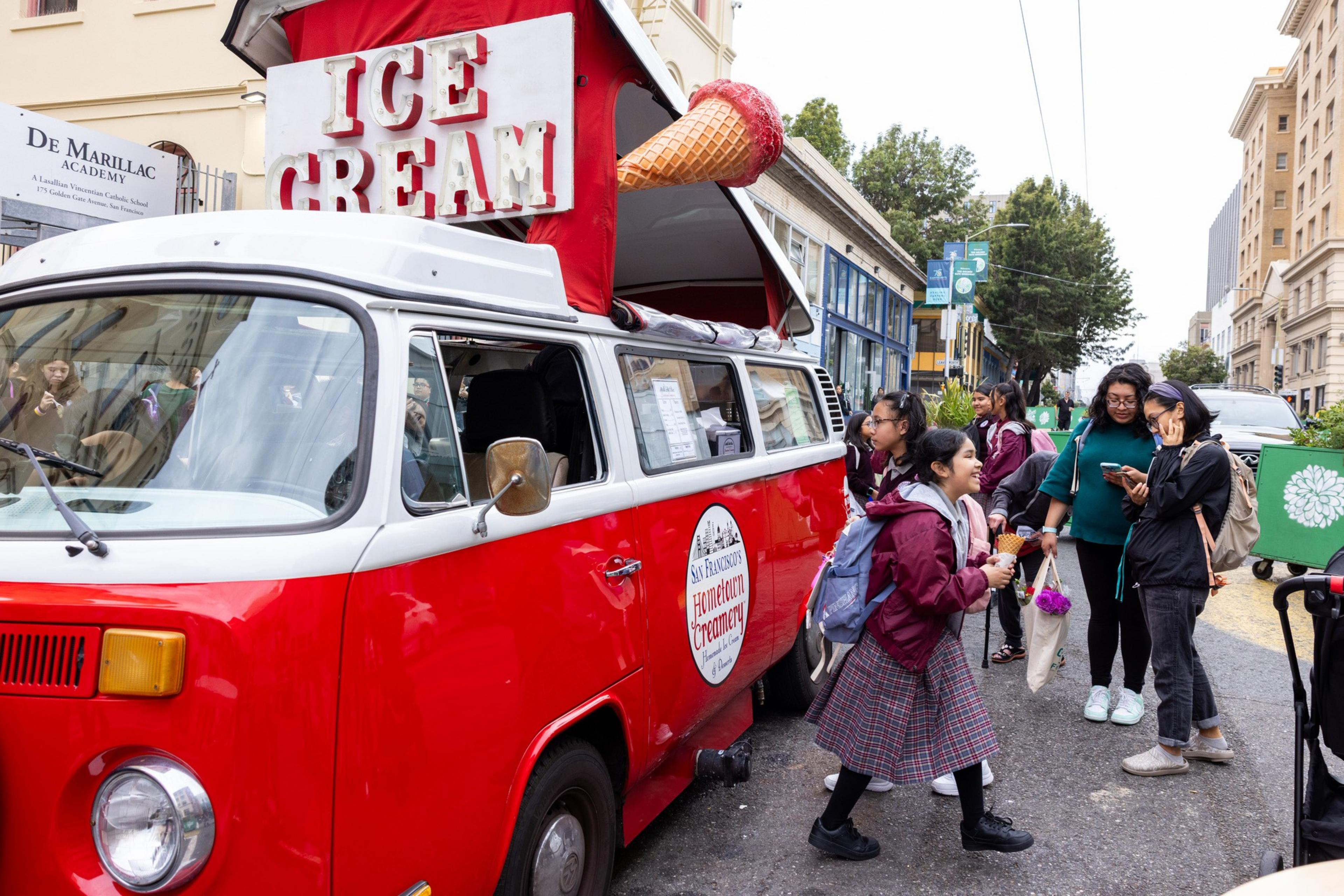 A red and white vintage ice cream van with a large ice cream cone sign on top serves customers on a city street with people gathered around it.