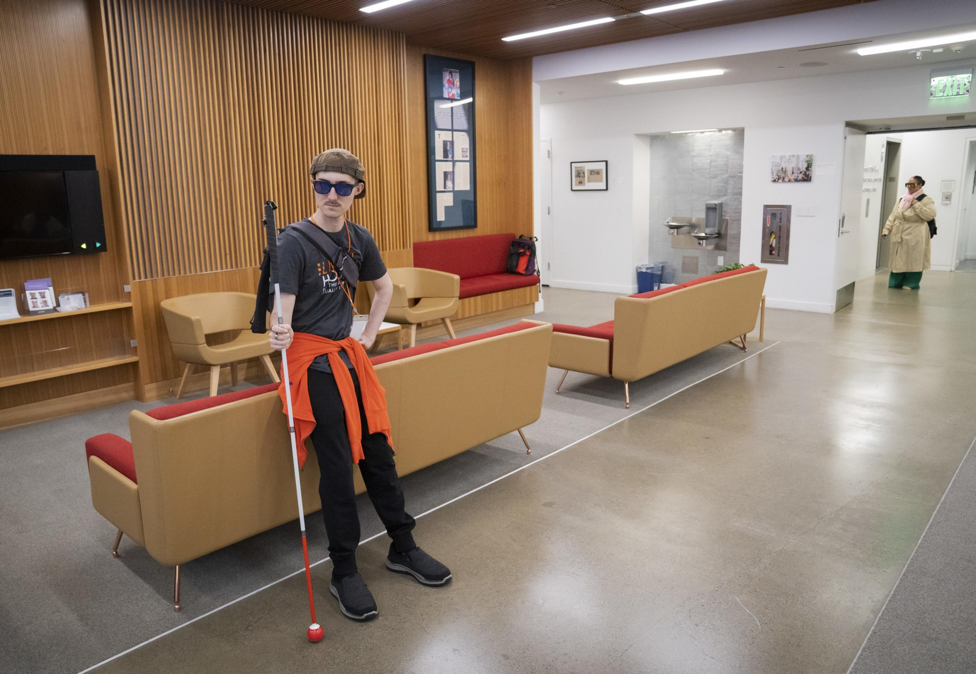 A person in sunglasses and a cap, holding a white cane, stands in a modern lounge with tan and red seating, wood panel wall. Another person waits for an elevator in a lobby area.