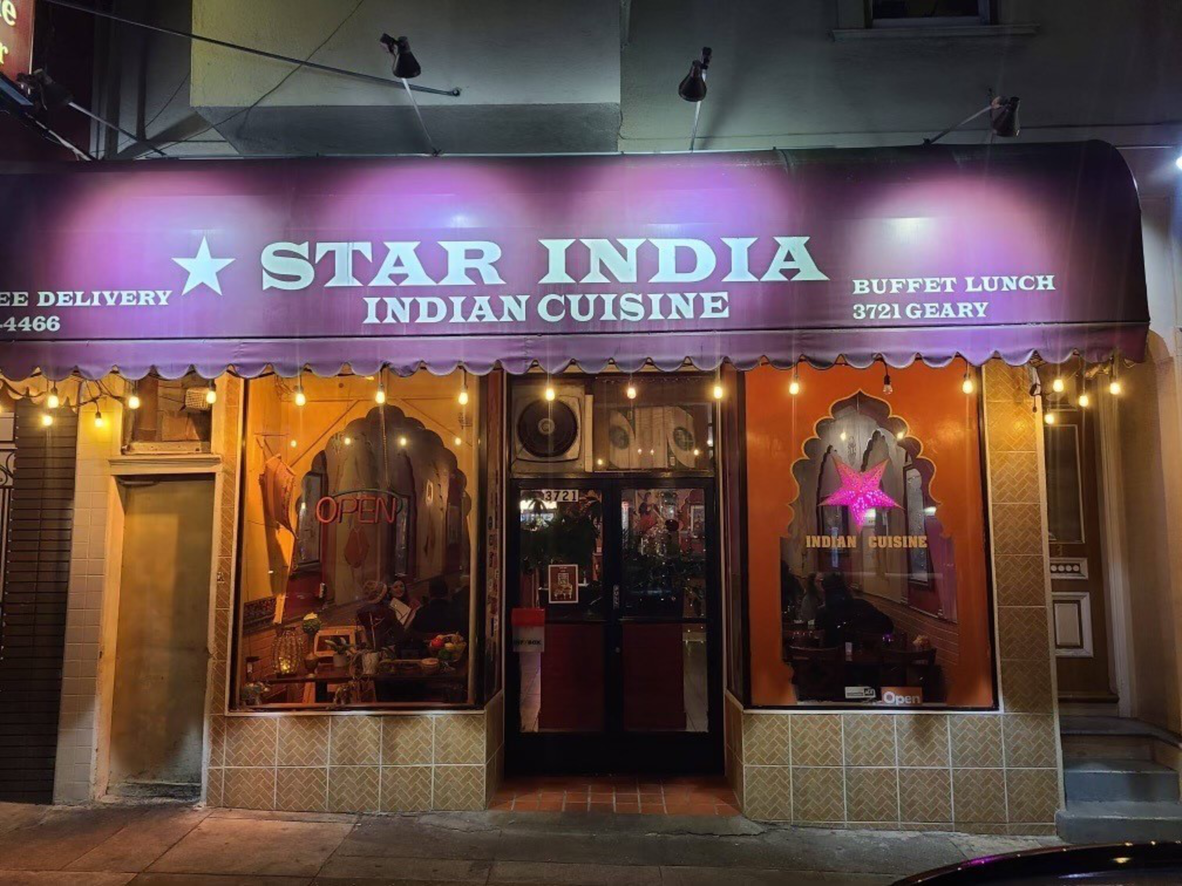 A purple awning with "Star India Indian Cuisine" in white letters covers a warmly lit restaurant entrance with two windows displaying decorations and signs.