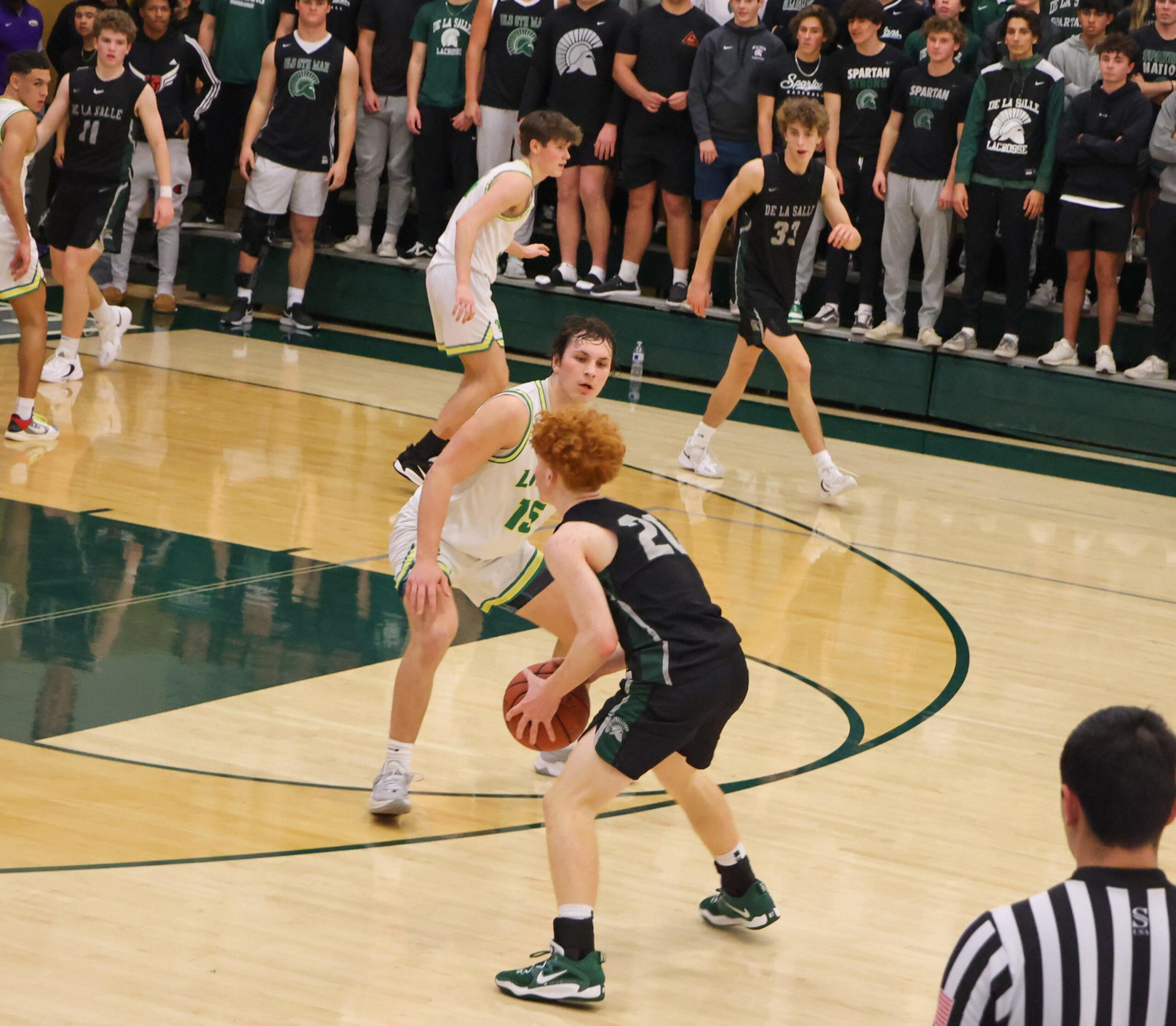 A basketball player in black dribbles while being closely guarded by a player in white on a gym court, with spectators and teammates watching behind.