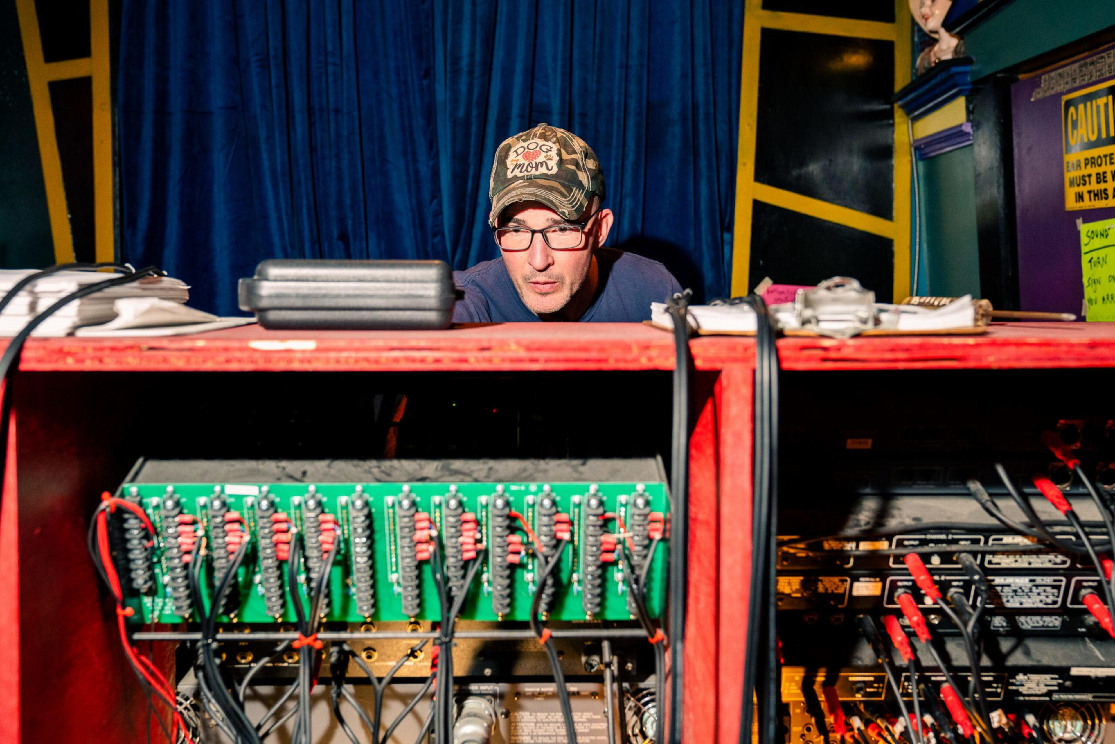 A man wearing glasses and a camo "Dog Mom" hat is working behind a red equipment rack filled with circuit boards and cables.